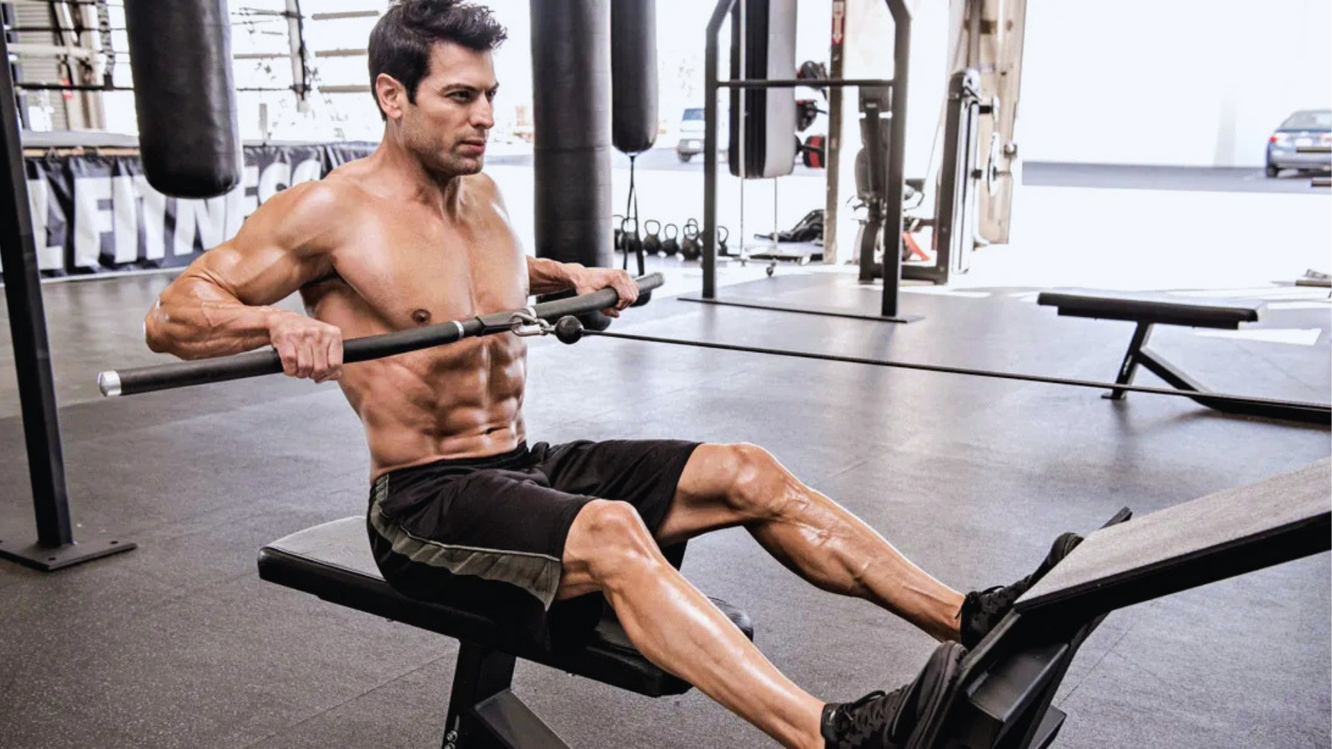 A muscular man works out on a rowing machine in a gym, showing focus and determination. He wears black shorts, and exercise equipment surrounds him.