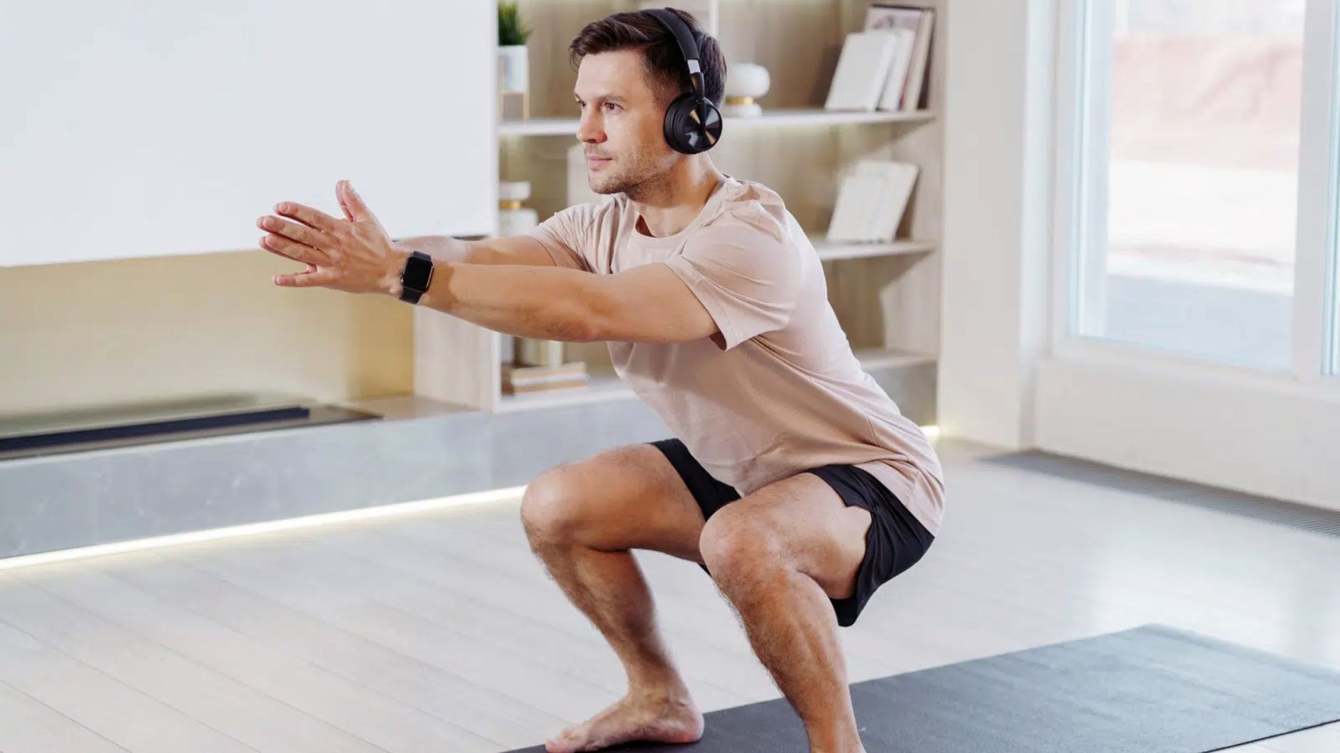 A man in a light shirt and shorts does a squat indoors, wearing headphones and an exercise watch. The room is bright with light wood flooring.