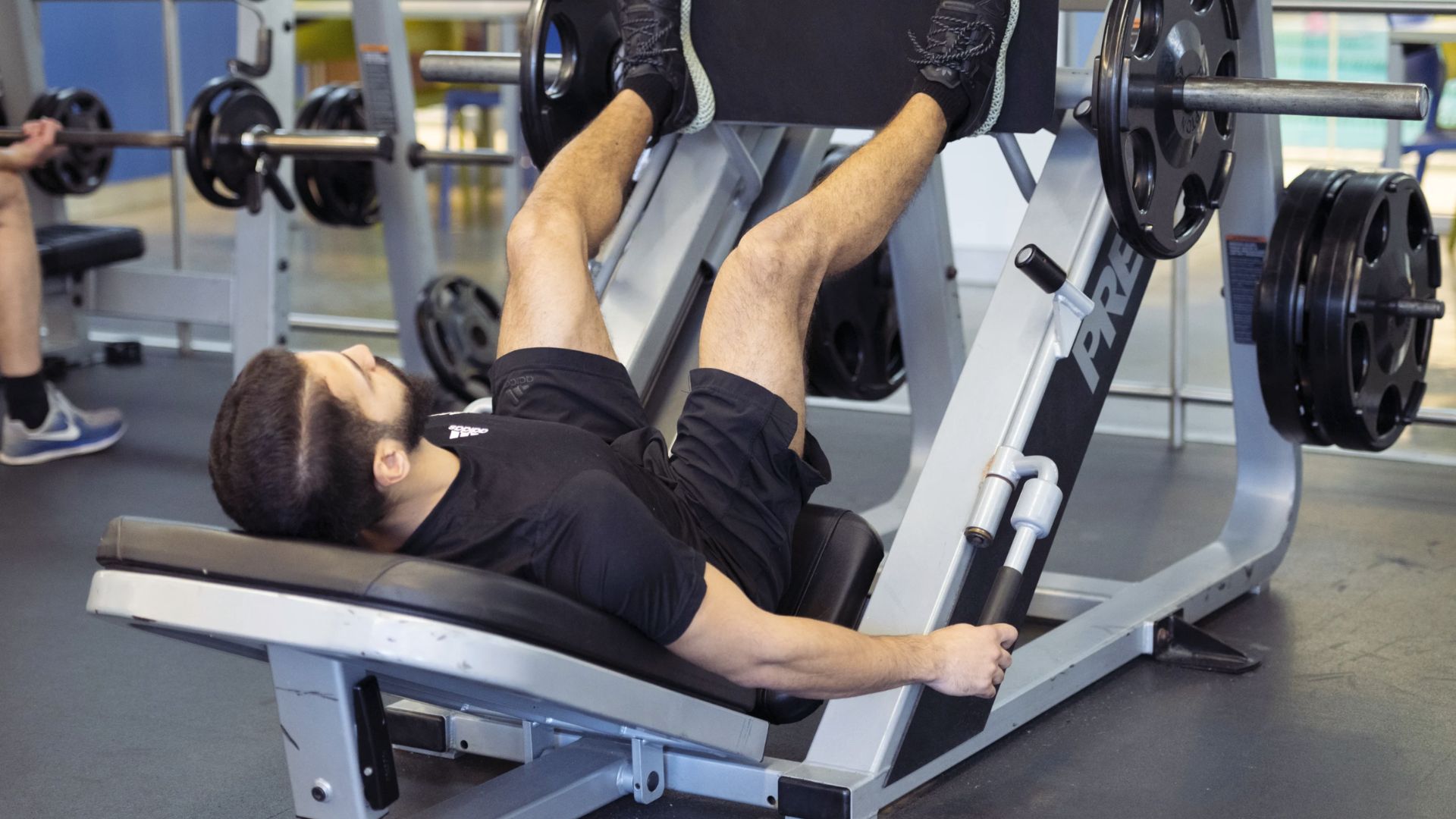 Man exercising on a leg press machine in a gym, wearing a black outfit. The focused expression conveys determination and effort in his workout.