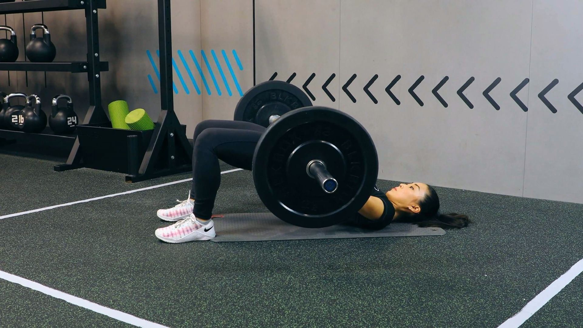 A woman performs a barbell hip thrust in a gym, with her back on a mat and feet flat on the floor. Kettlebells and weights are visible in the background.