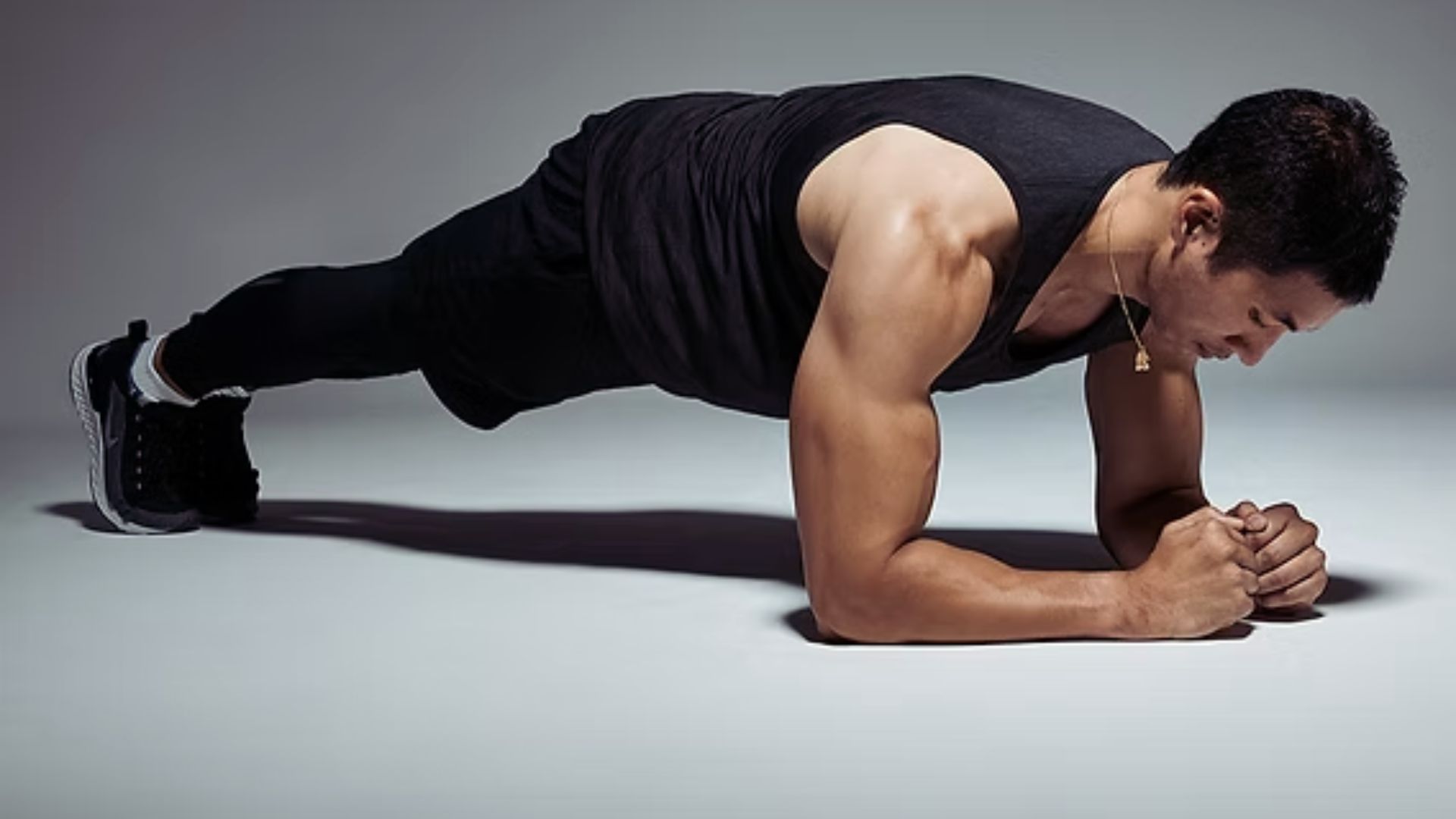 A man in a dark athletic outfit holds a plank position on a neutral background. His focused expression and muscular build convey strength and determination.