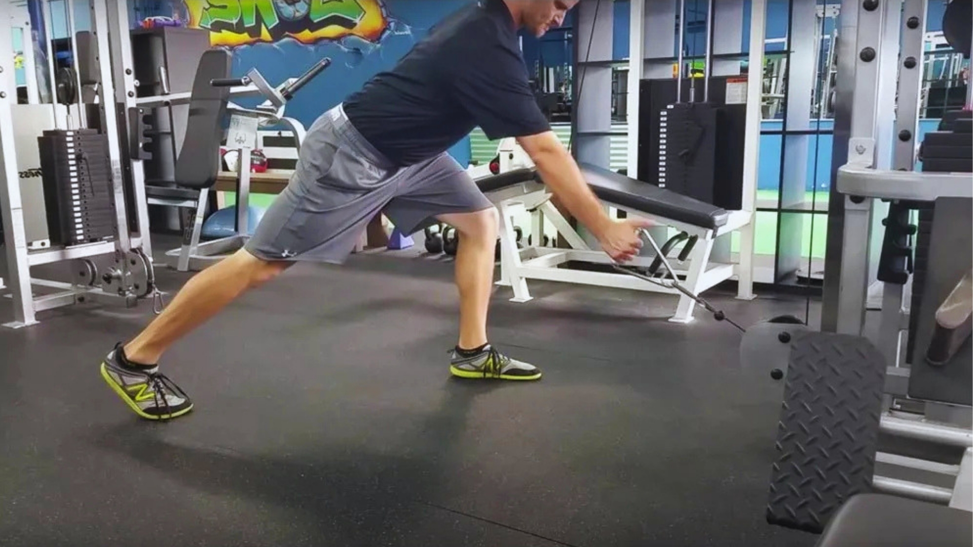 A person in a gym uses a cable machine, lunging forward in athletic shoes on a black floor. Bright graffiti art decorates the blue wall behind.