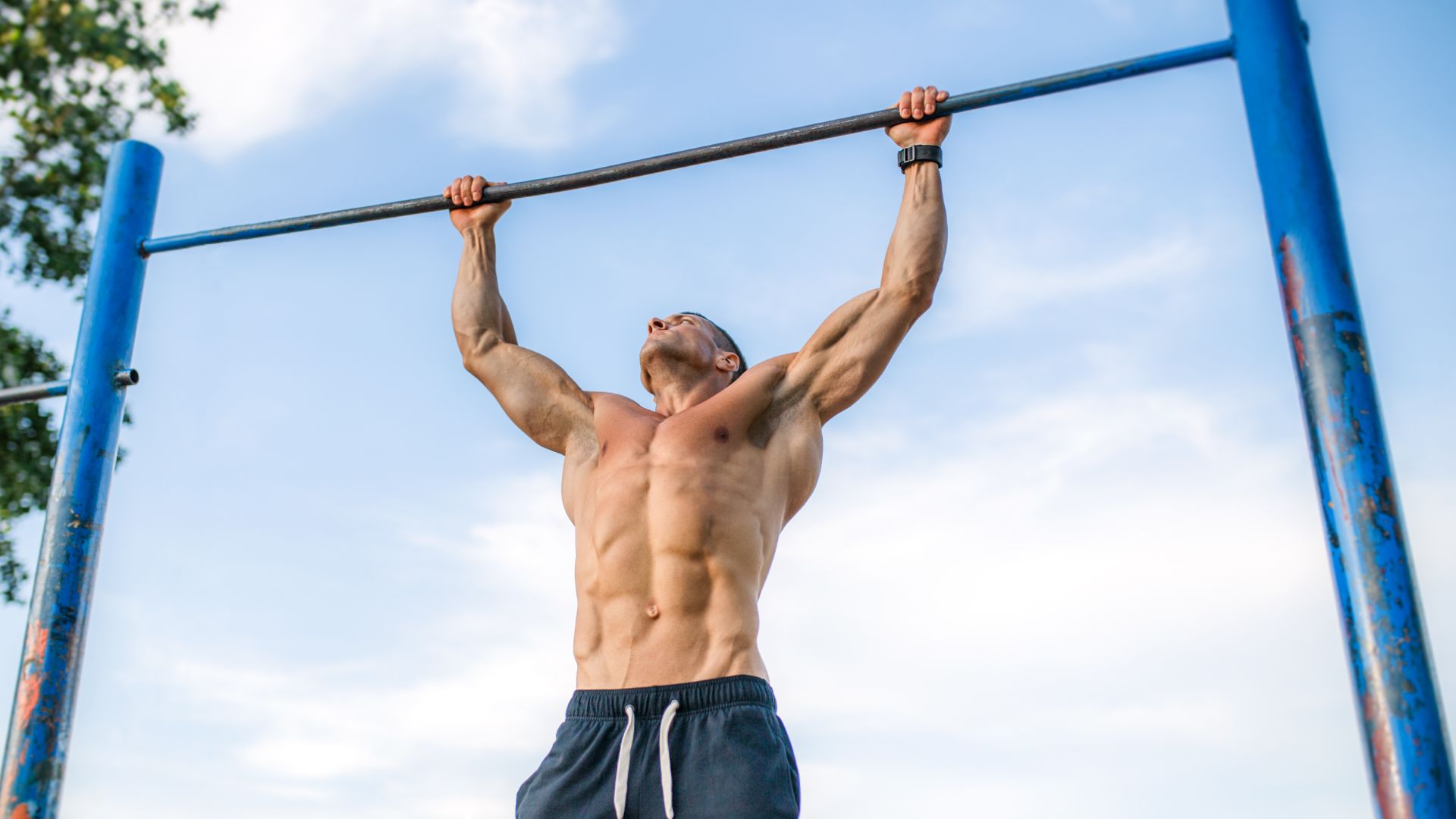 A muscular man performs a pull-up on a blue bar outdoors, displaying strength and focus. The sky and trees are visible in the background.