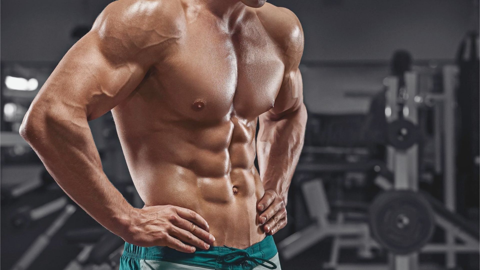 Muscular man with hands on hips, showcasing defined abs and arms. He's wearing shorts in a gym setting, conveying strength and determination.