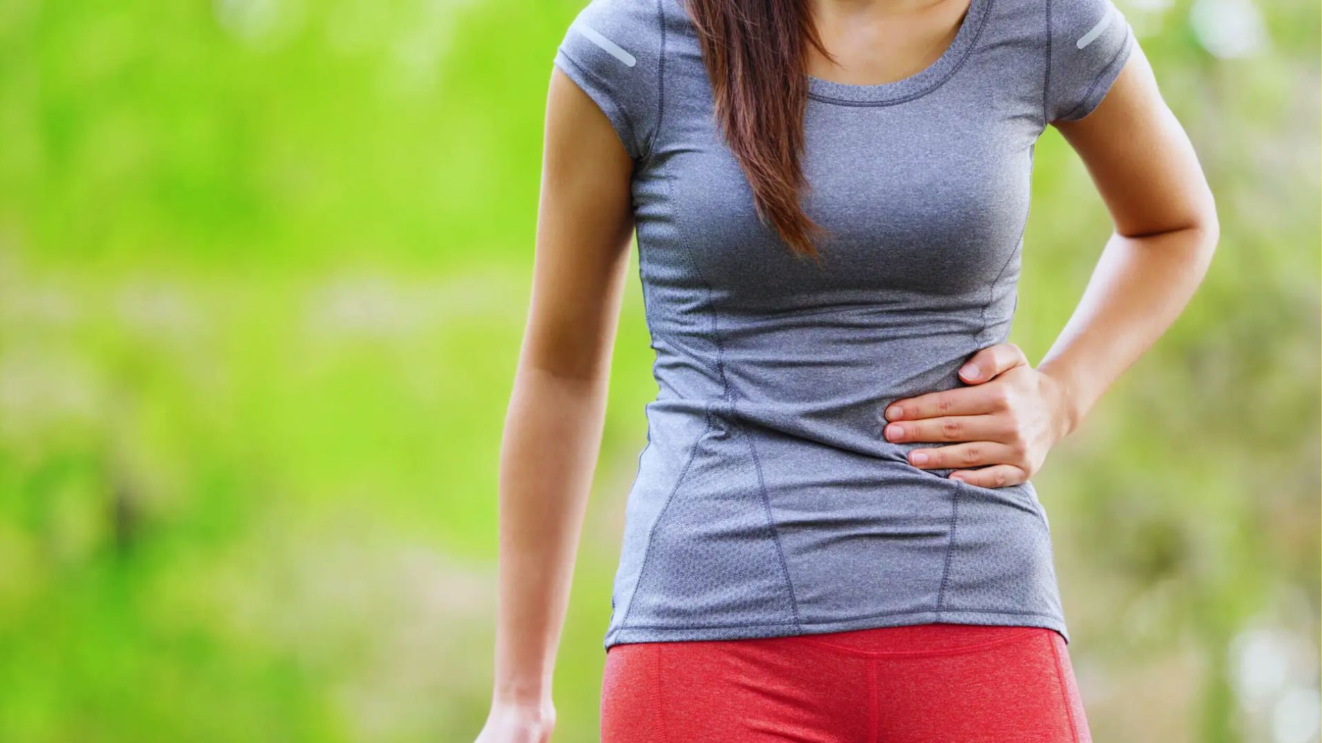 A woman in workout attire clutches her side, appearing to be in discomfort. She is outdoors, wearing a gray top and red pants, with a blurred green background.