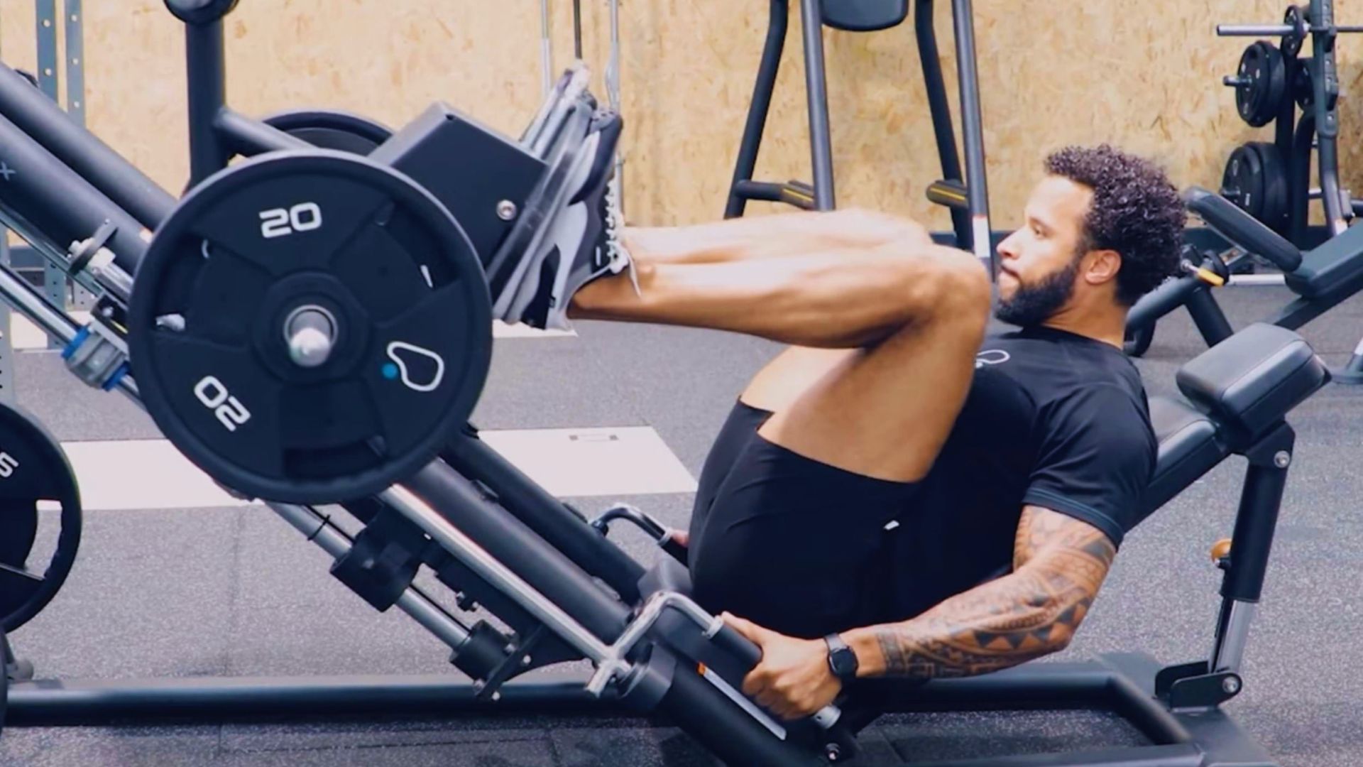 A man in a gym is performing a leg press exercise on a machine, wearing a black shirt and shorts. He appears focused and determined.