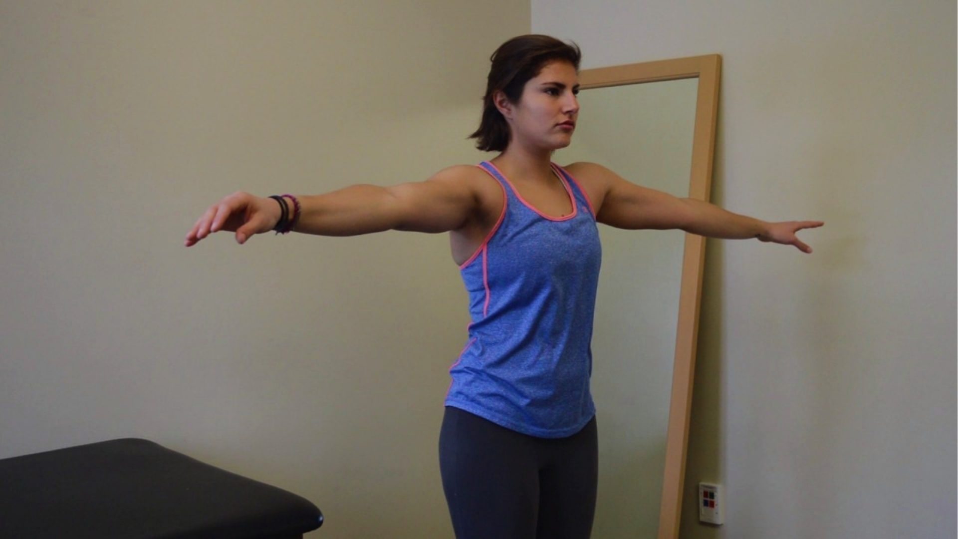 A woman in workout attire stands with arms outstretched, parallel to the floor. She appears focused, performing a balance or fitness exercise in a plain room.