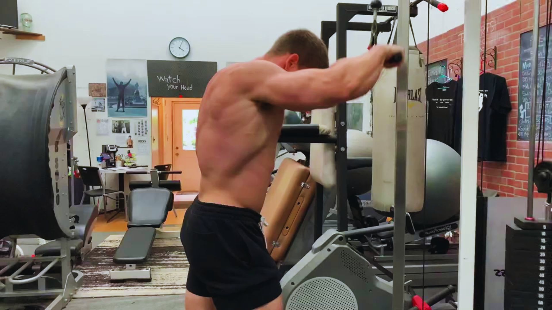 A muscular person in black shorts exercises on a cable machine in a gym. The room has fitness equipment, motivational posters, and a relaxed ambiance.