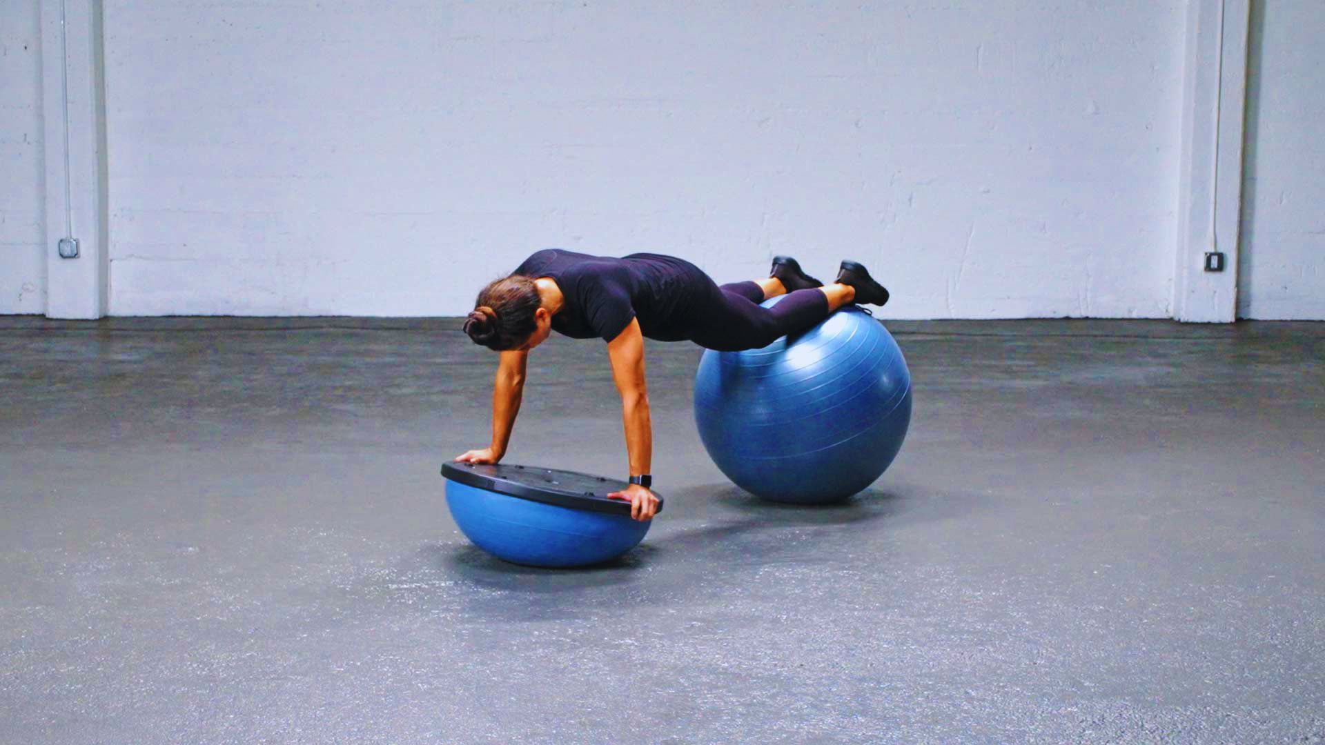 A person in athletic wear balances on a blue stability ball with their hands on a Bosu ball, performing a plank in a spacious, minimalist gym.