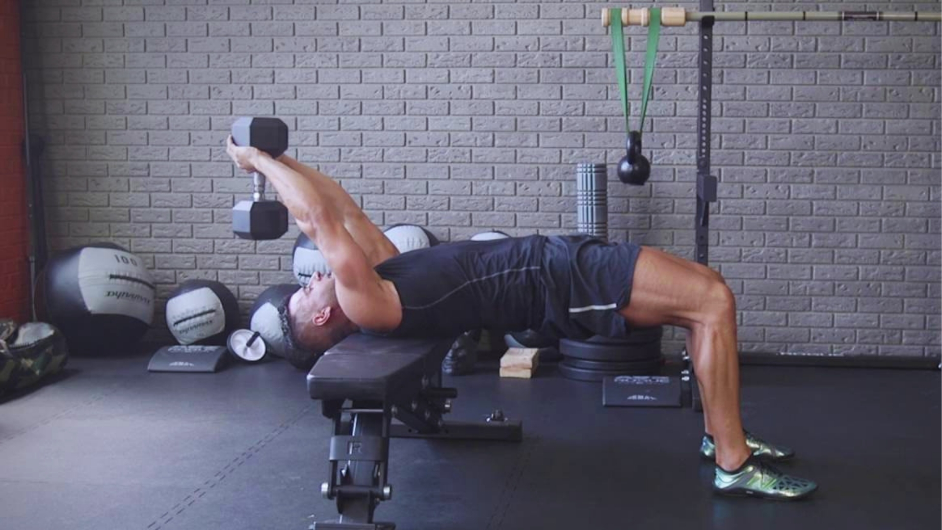 Man in athletic wear performs a dumbbell pullover on a bench in a gym. The setting includes a textured wall, exercise balls, and resistance bands, conveying strength and focus.