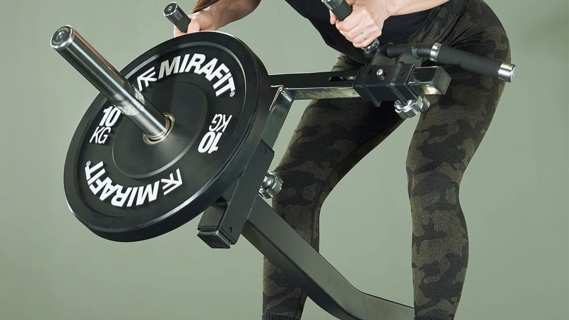A person in camo leggings is using a Mirafit landmine attachment with a 10 kg weight plate. The setup is designed for strength training exercises.