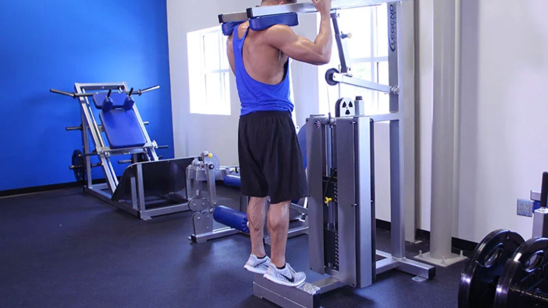A man in a blue tank top and black shorts performs a calf raise on a weight machine in a gym, conveying focus and strength in a well-lit room.