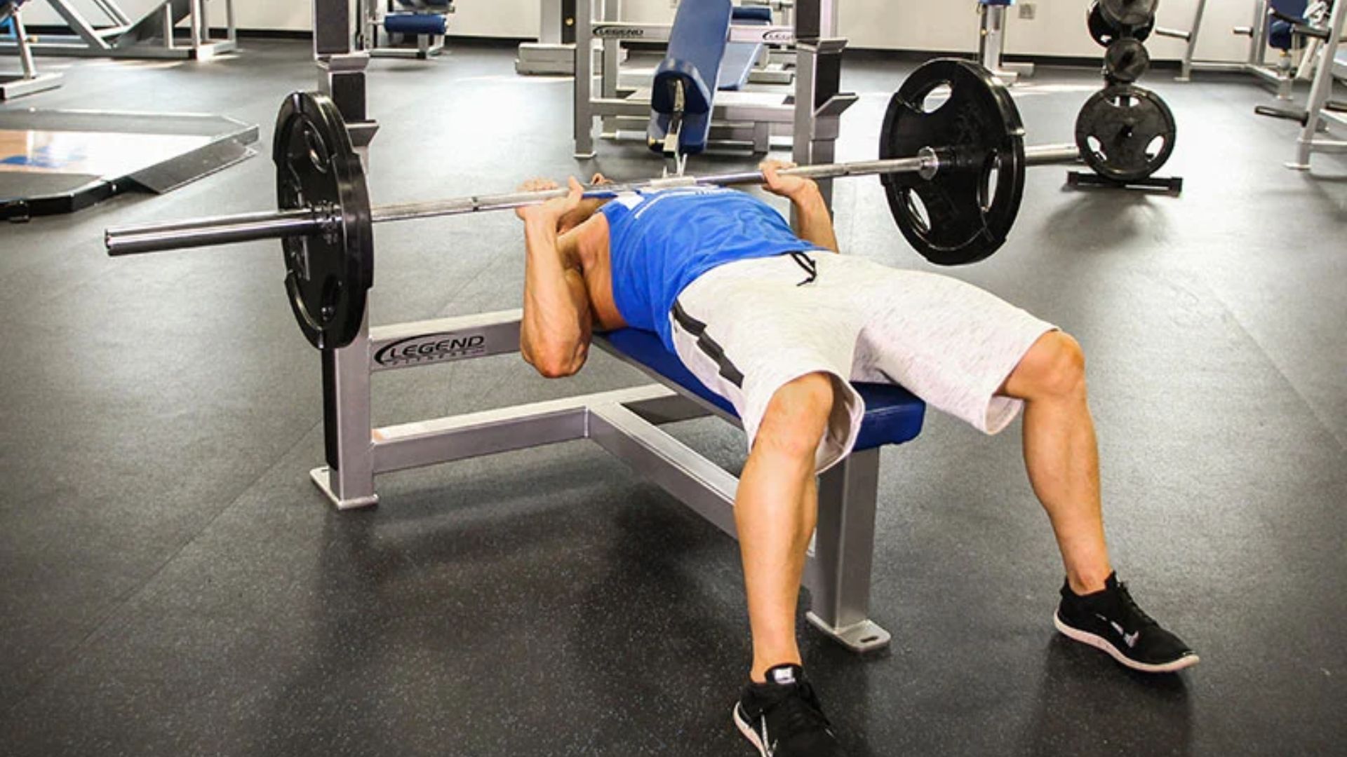 Man in blue tank top and shorts bench pressing in a gym, focusing on strength training with weights on a barbell.