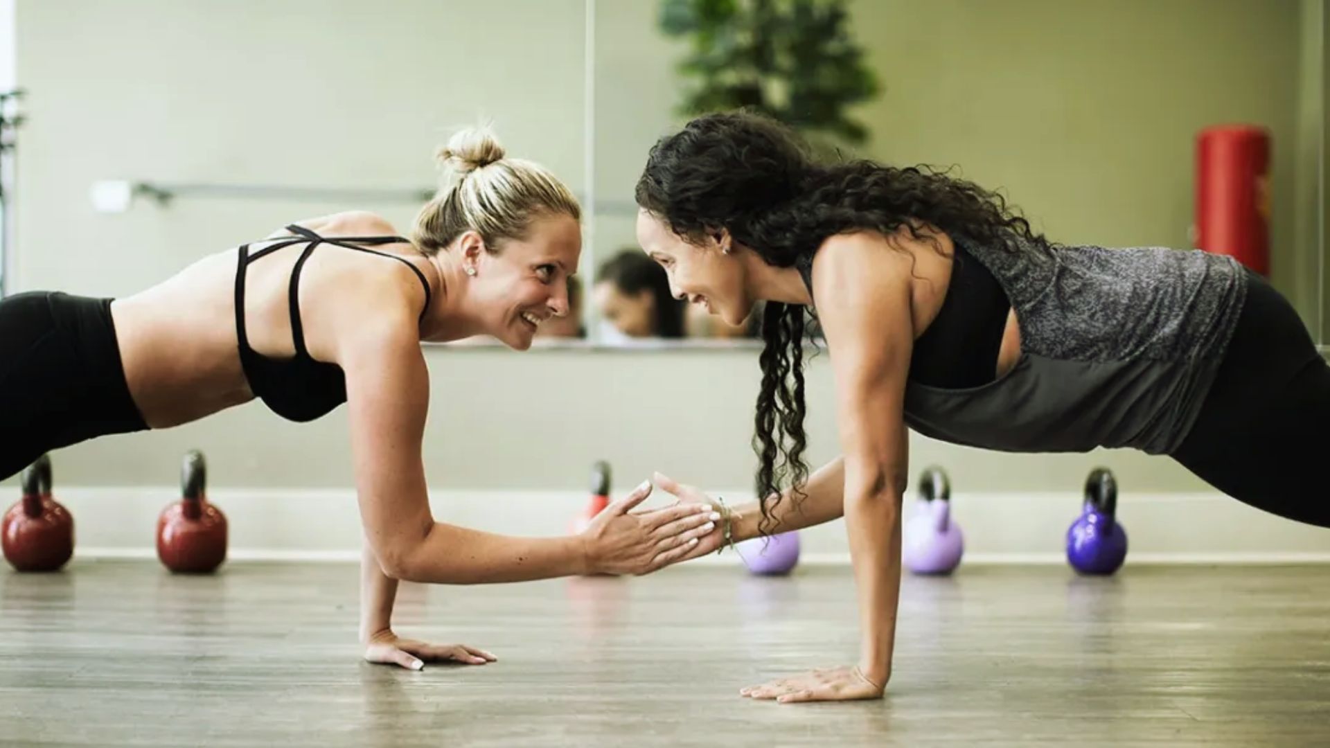 Two women in workout gear hold a plank pose, smiling and high-fiving in a gym. Red and purple kettlebells are in the background, with a lively, supportive atmosphere.
