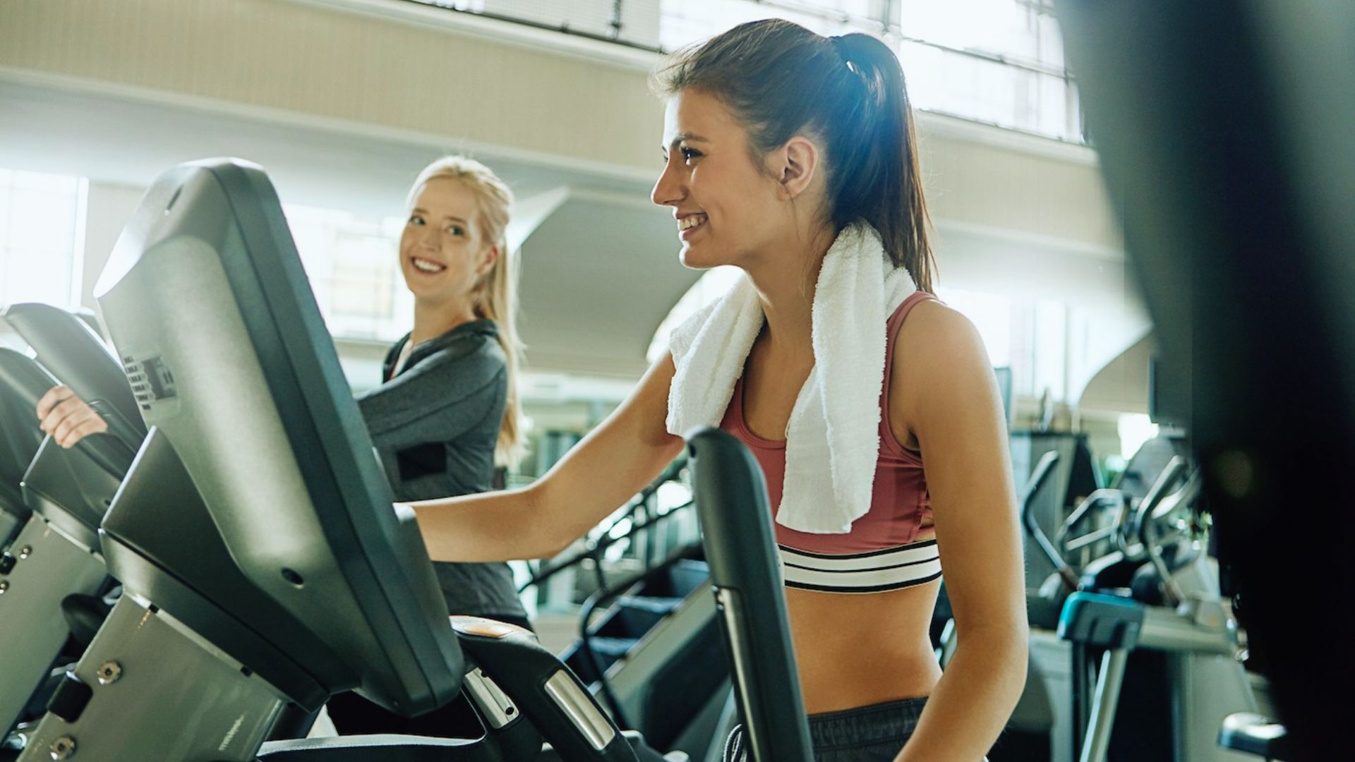 Two women exercise on treadmills in a bright gym. The woman in front smiles, with a towel around her neck, conveying a positive and energetic mood.