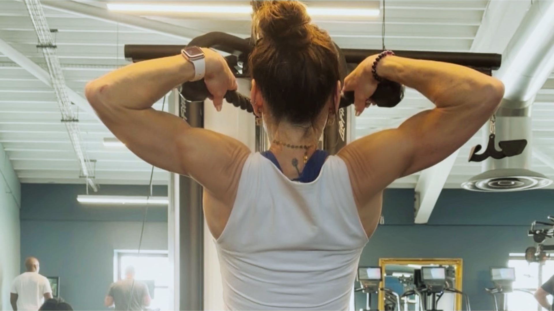 A woman in a white tank top works out her back muscles on a cable machine in a gym. The setting is bright and modern, with people exercising in the background.