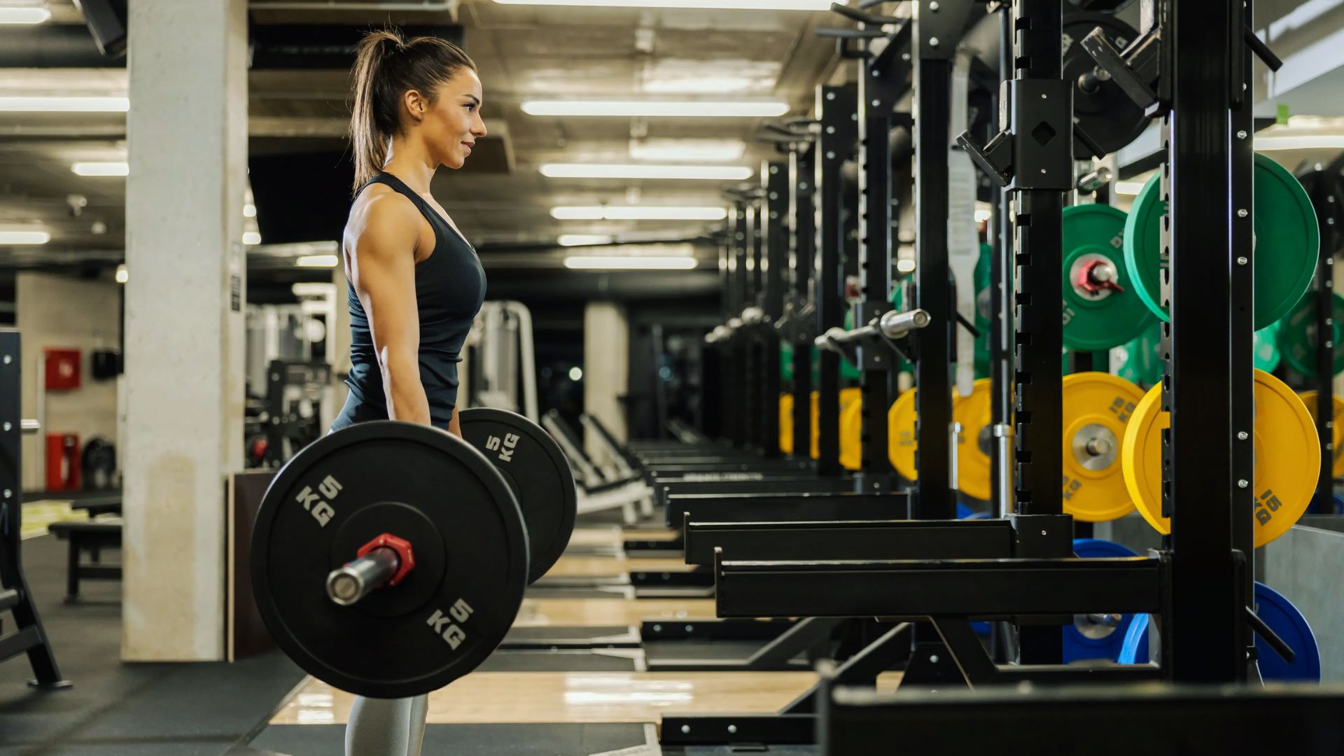 A woman in athletic wear performs a deadlift in a gym, surrounded by colorful weight plates. The setting is modern, focused, and energetic.