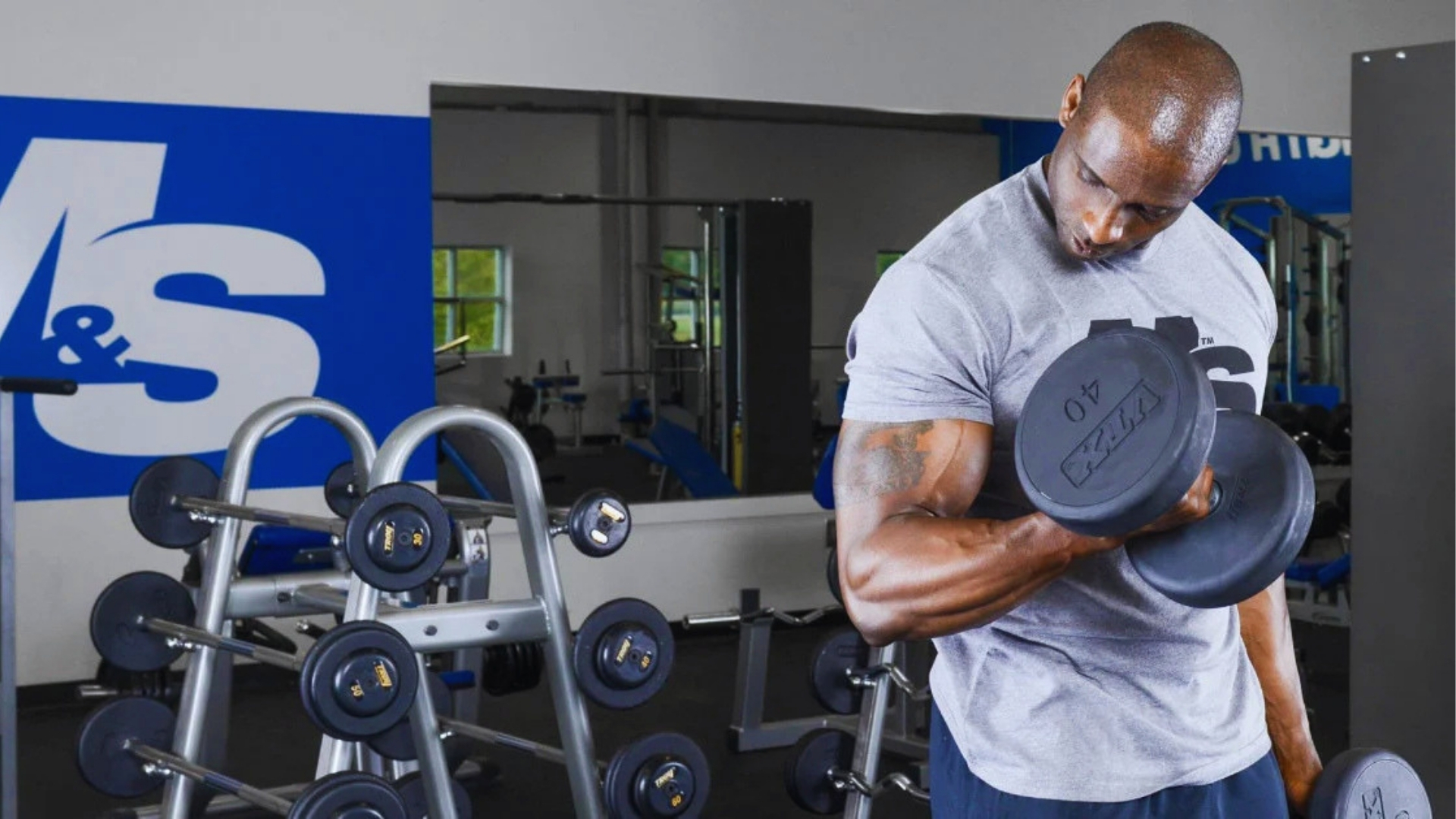 A muscular man in a gray t-shirt lifts a 40-pound dumbbell in a gym. Rows of weights are visible, with gym equipment and mirrors in the background.