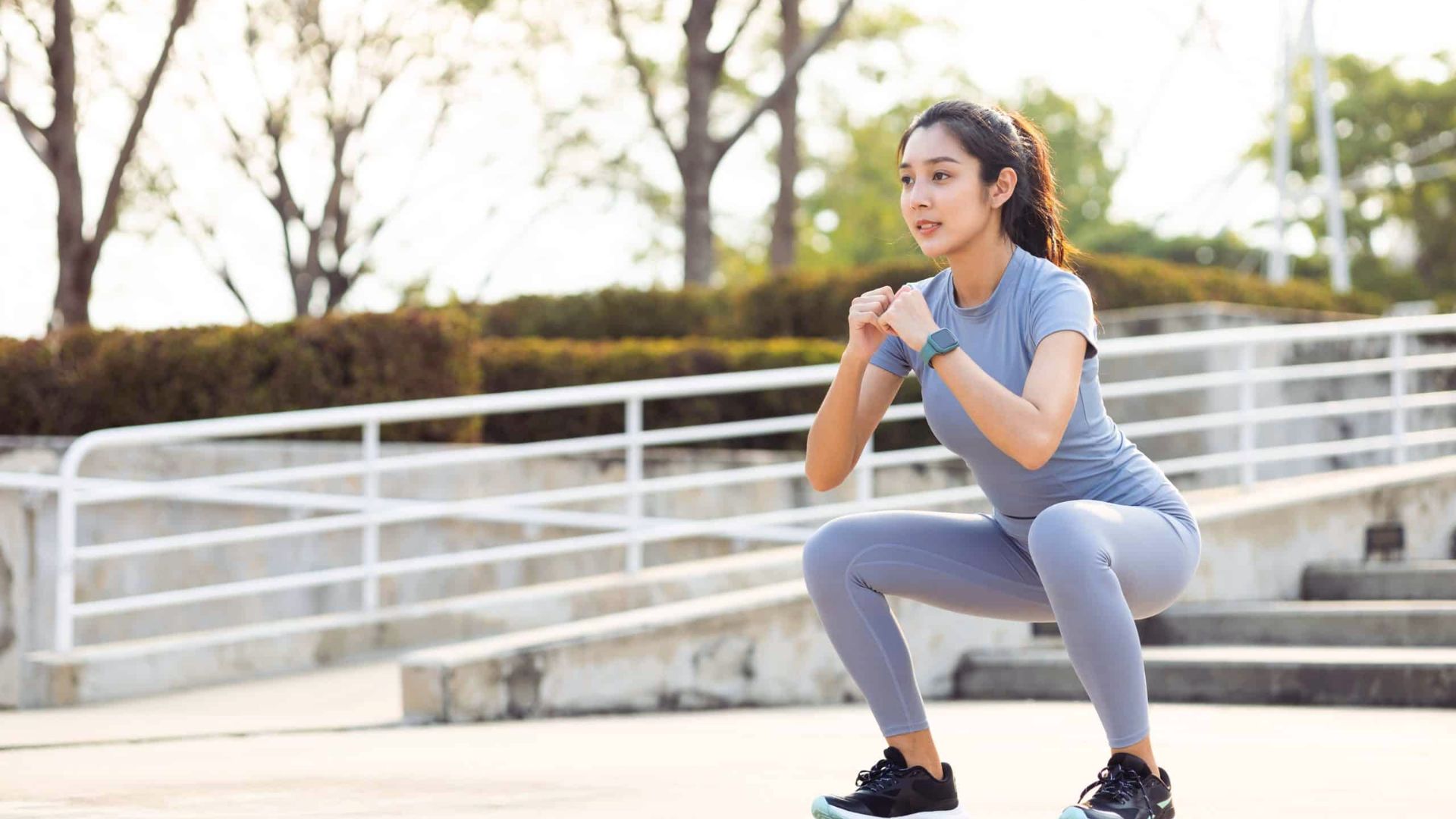 A woman in athletic wear performs squats outdoors, surrounded by trees and railings. She appears focused and determined, exercising in a sunny park.