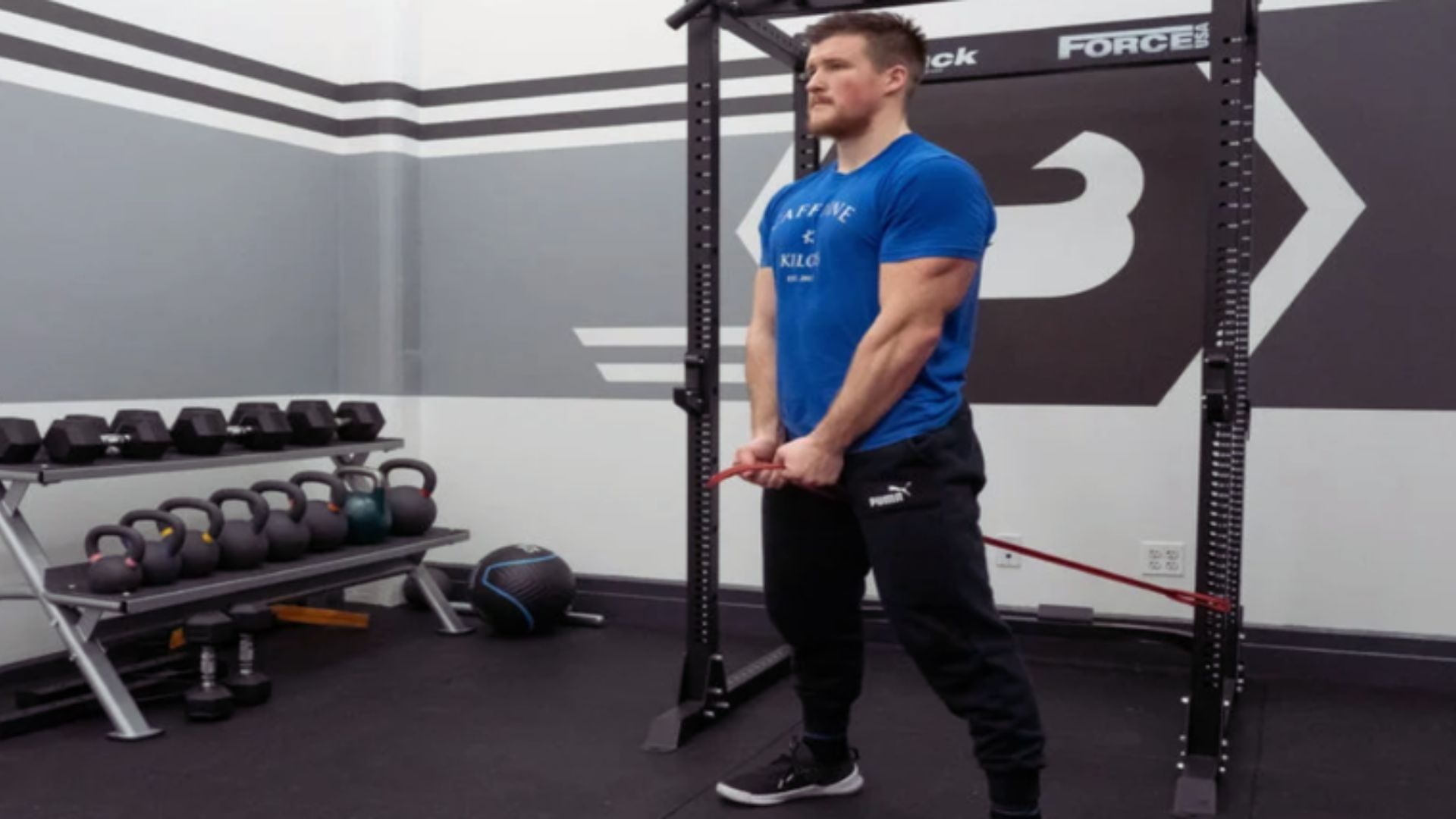 A man exercises with a resistance band in a gym. He wears a blue shirt and black pants, surrounded by weights and fitness equipment, conveying focus.