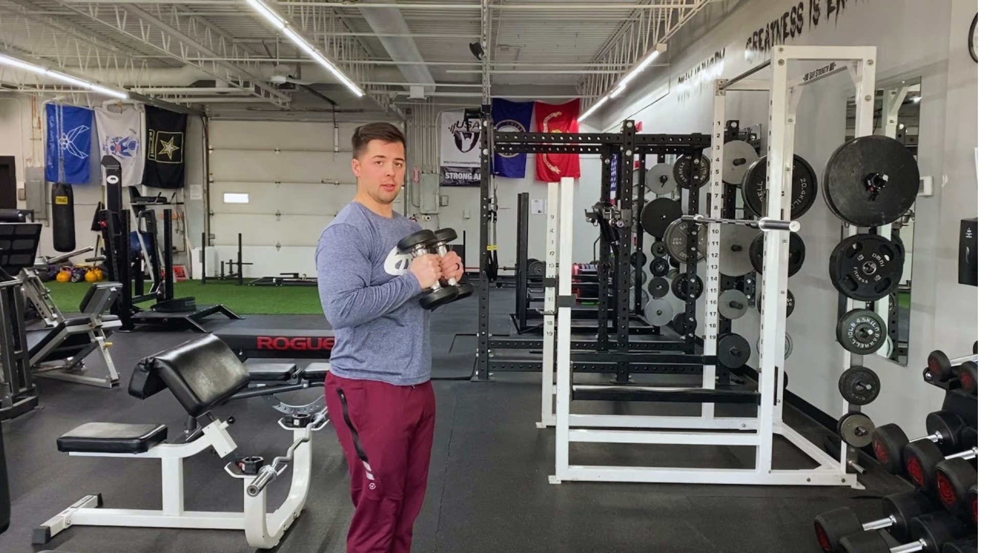 A man in maroon pants and a gray long-sleeve shirt holds a kettlebell in a bright gym with weightlifting equipment and flags on the walls.