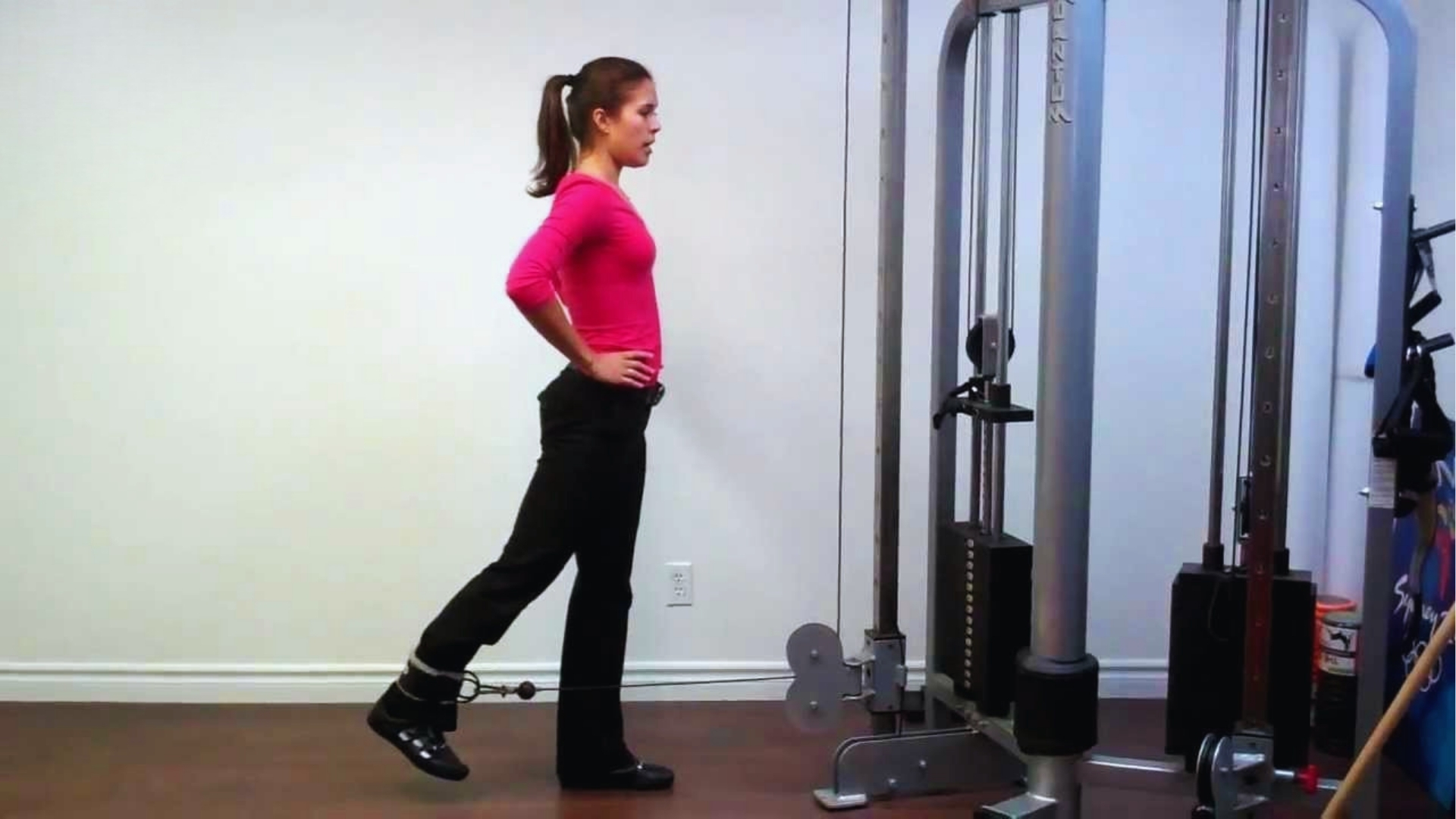A woman in a pink top and black pants performs a cable leg exercise at the gym. She stands sideways, focusing intently, with hands on hips.