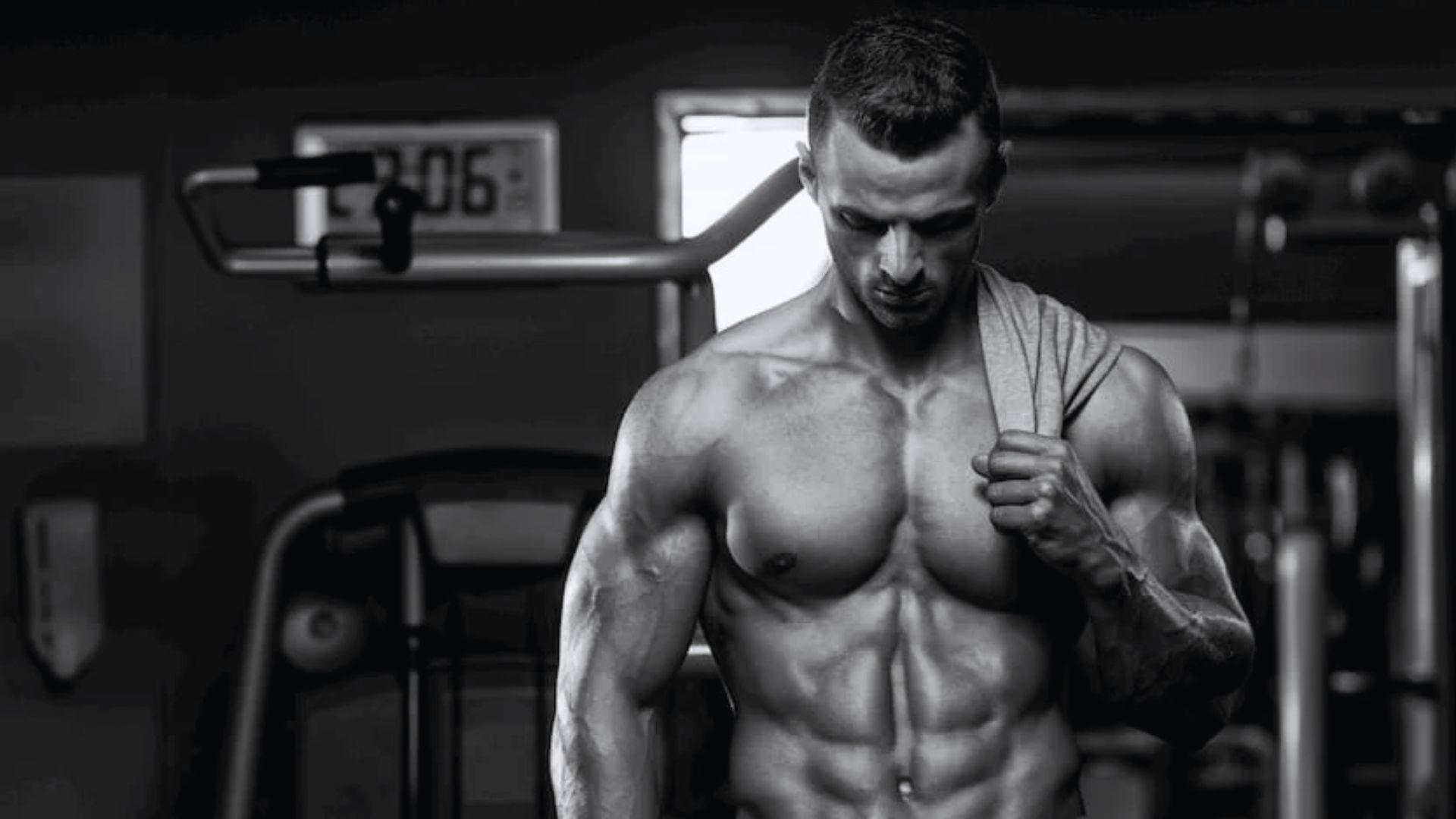 Muscular man in a gym, shirt draped over shoulder, gazing down. Black and white tones highlight physique and reflect determination. Fitness equipment in background.