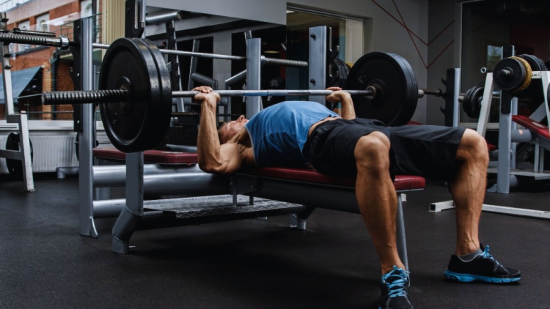 A man in a blue tank top and black shorts performs a bench press in a gym, focusing intently. The room is equipped with various weights and machines.
