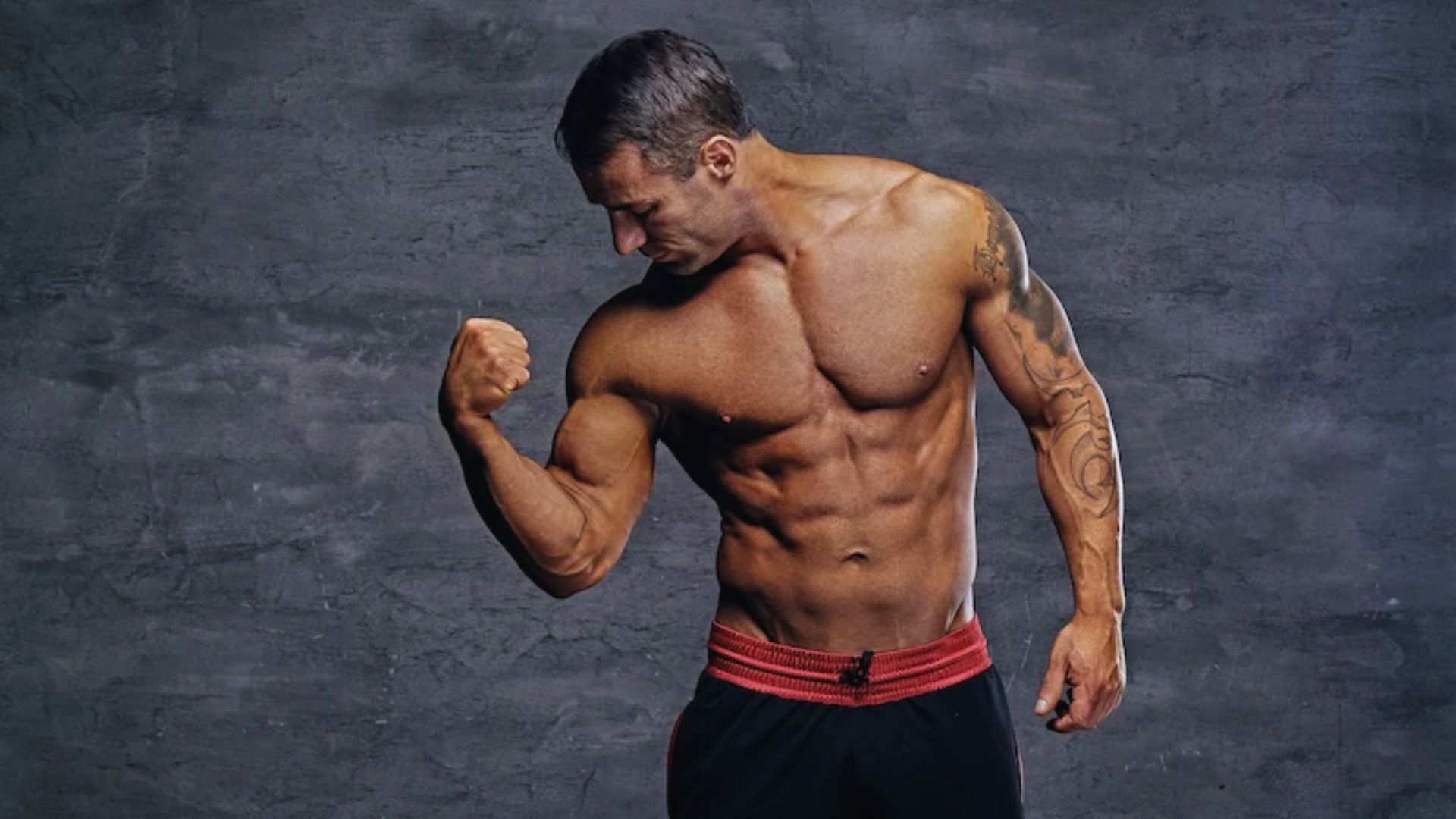 Muscular man with tattoos flexes his bicep while wearing black shorts with a red waistband. He stands against a dark textured background.