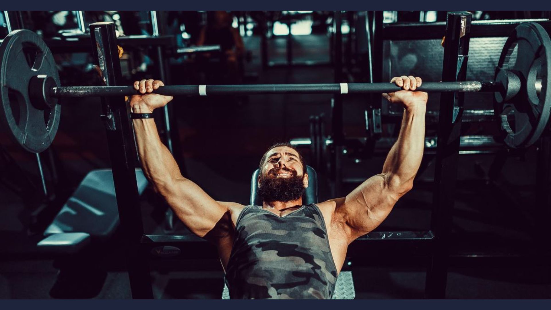 A bearded man in a camo tank top bench presses a weighted barbell in a dimly lit gym, displaying focus and strength. Equipment surrounds him in the background.