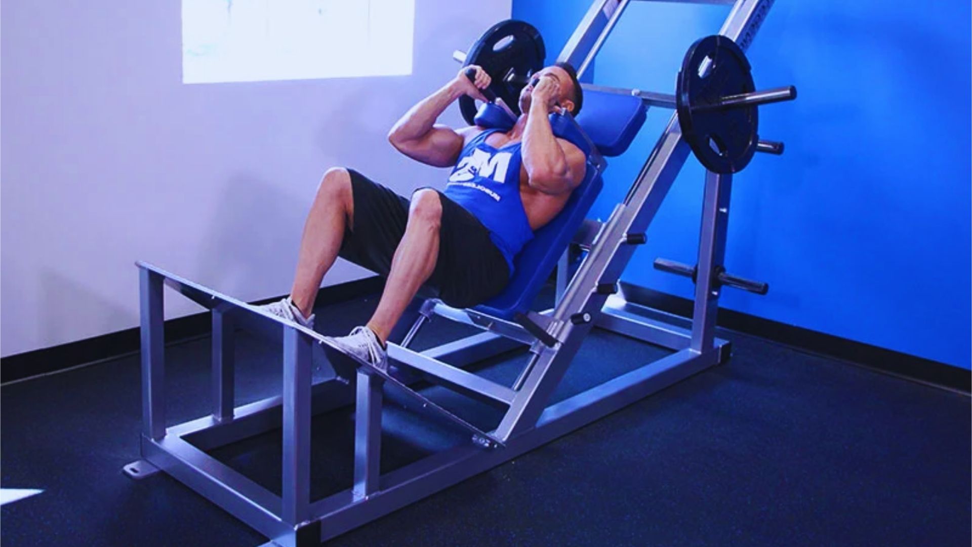 A man in a blue tank top is doing a leg press on a hack squat machine. He is in a gym with blue walls, focused and exerting effort.