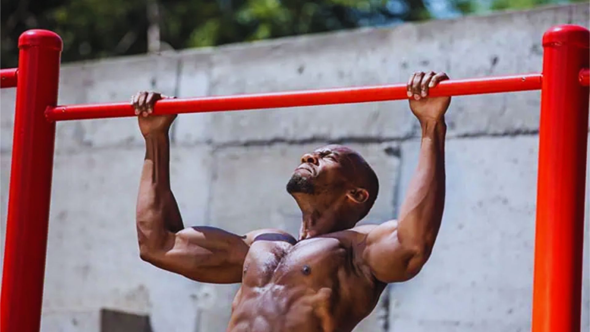 A muscular man performs a pull-up on a vibrant red bar, displaying strength and determination. A concrete wall and trees form the outdoor background.