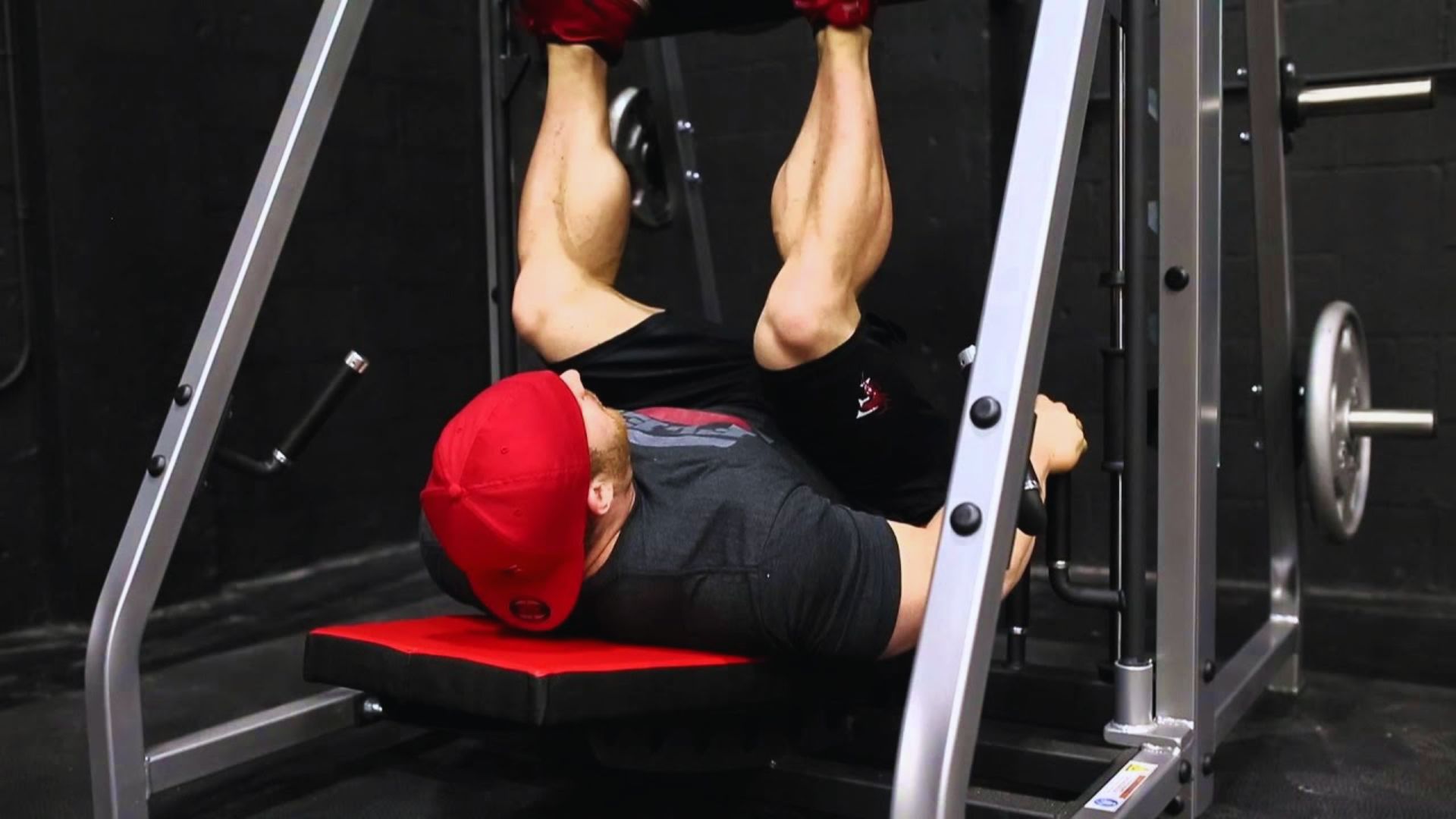 A person in a red cap and black gym attire performs leg presses on a weight machine. The setting is a dimly lit gym, conveying focus and determination.