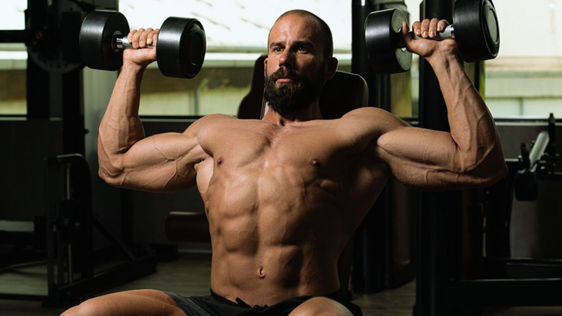 A muscular man with a beard is lifting dumbbells in a gym, showcasing strength and focus. The gym is dimly lit, highlighting his physique.