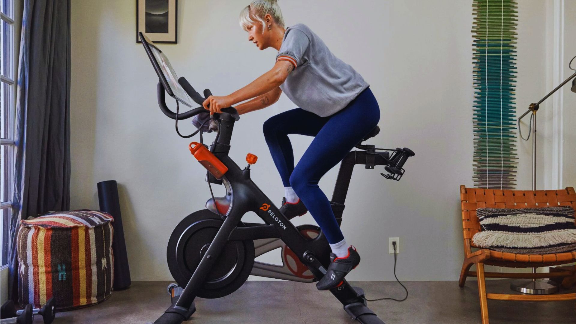 A person exercises on a Peloton bike at home, looking forward with focus. The room is cozy, featuring a woven chair, striped cushion, and colorful wall art.