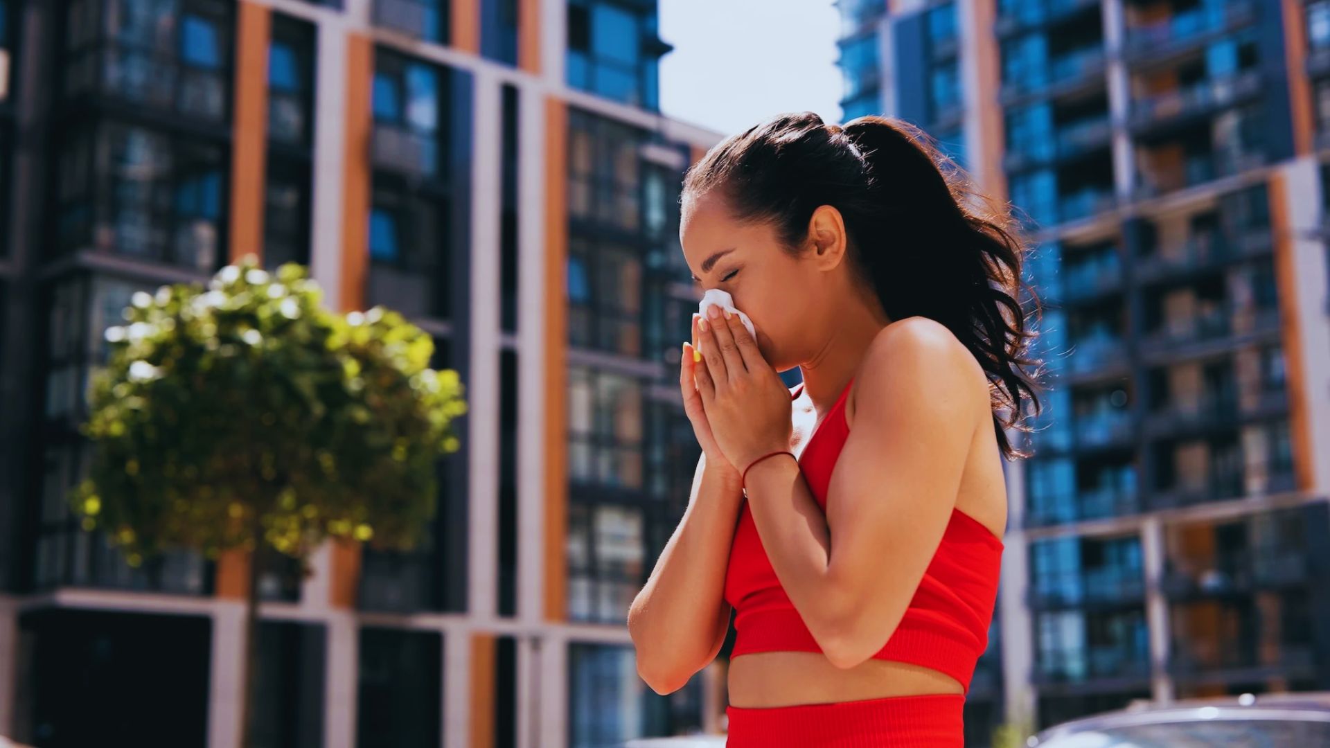 A woman in a red workout outfit sneezes into a tissue outdoors. She stands in front of modern buildings and trees, appearing slightly uncomfortable.