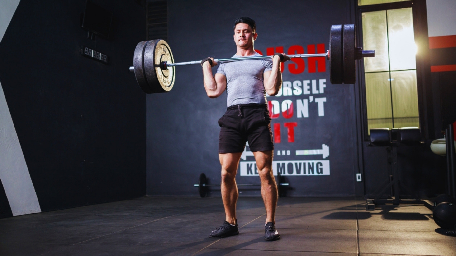 A man lifts a barbell in a gym, focused and determined. Motivational text on the wall reads "Push Yourself, Don't Quit, Keep Moving."