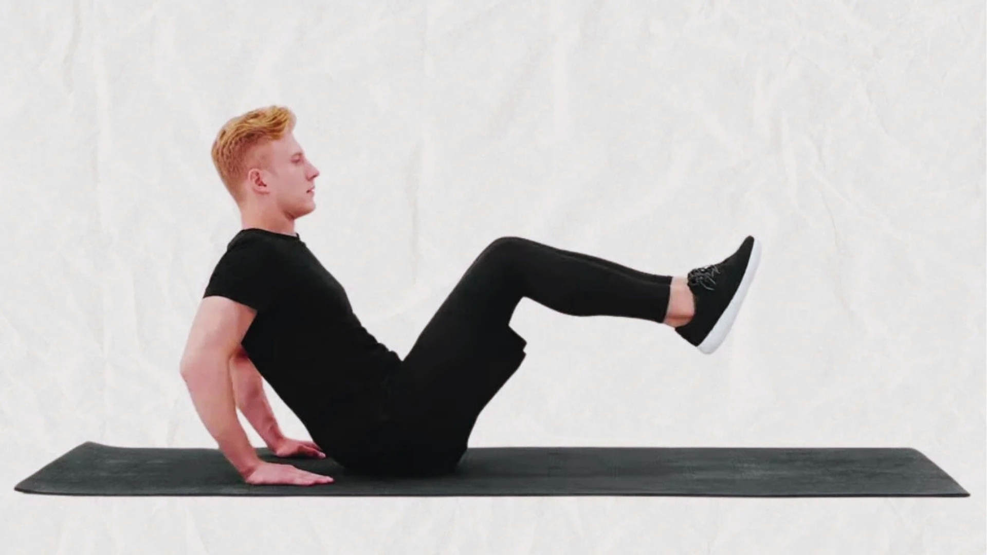 Person in a black outfit performing a core exercise on a mat with legs lifted and bent, hands on the ground for support, on a white background.