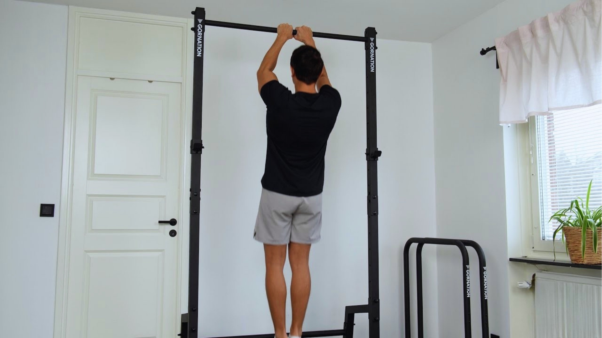 A man in a black shirt and gray shorts performs a pull-up on an indoor bar. The room is bright, with a potted plant on the windowsill.