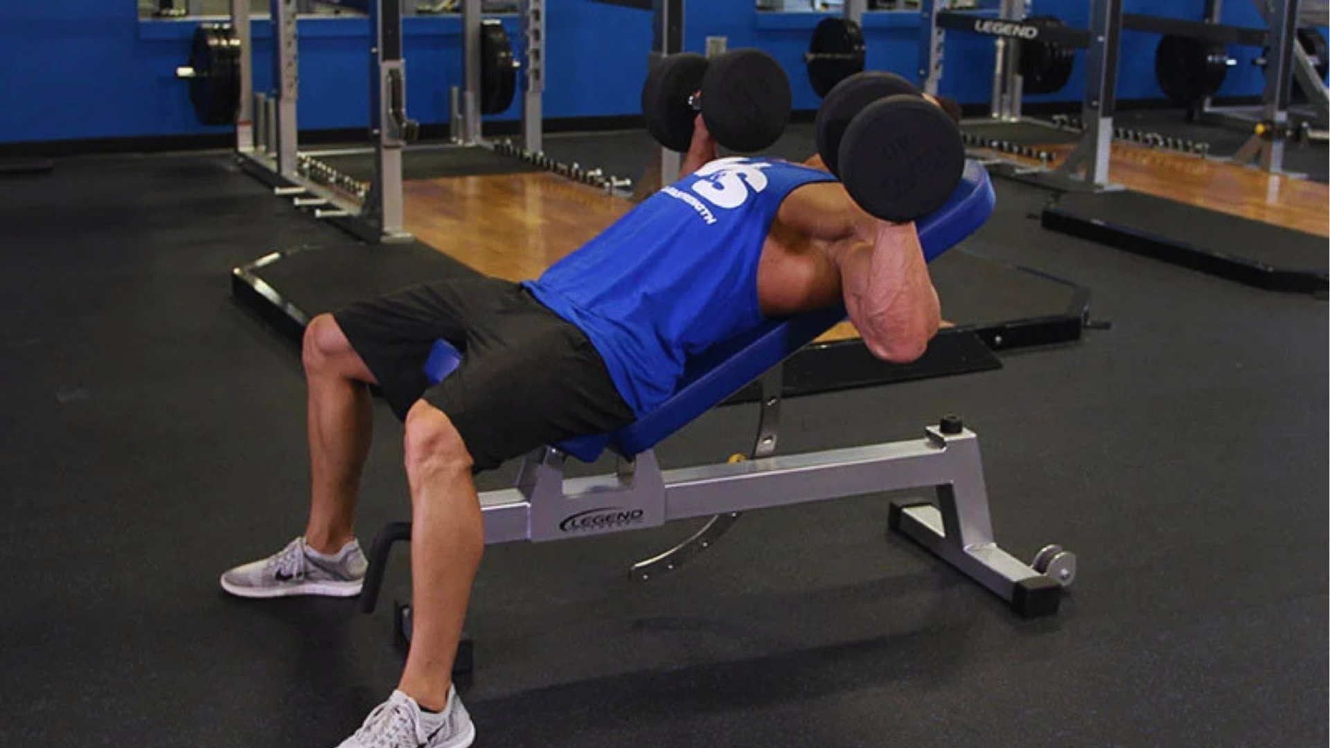 A man in a blue tank top is performing an incline dumbbell press on a bench in a gym. The setting is focused and energetic, highlighting strength training.