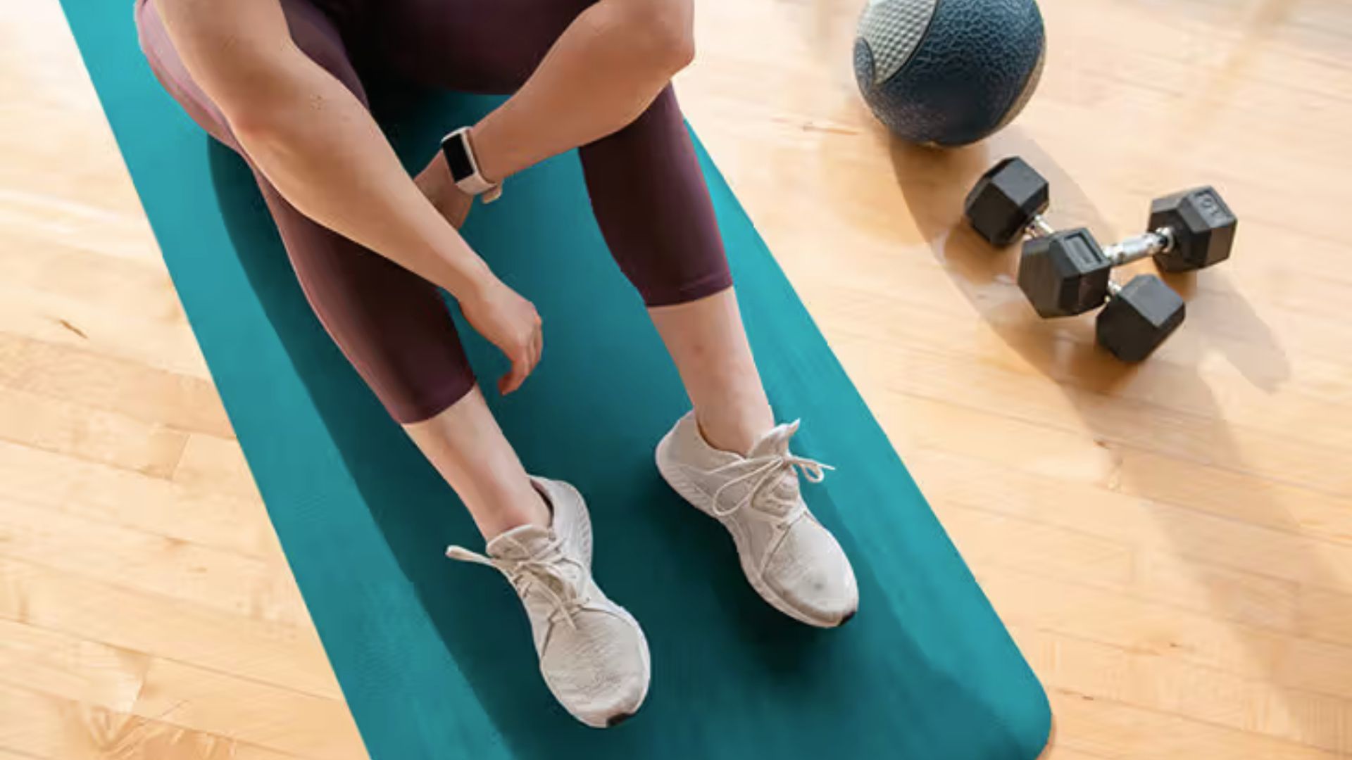Person sitting on a teal mat wearing workout gear and white sneakers, adjusting a smartwatch. Nearby are dumbbells and a medicine ball on a wooden floor.