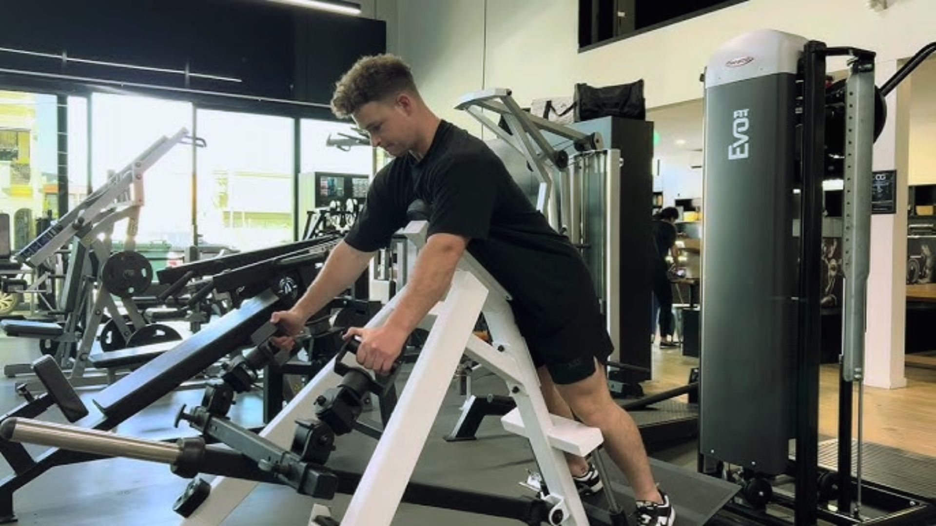 A person in a black workout outfit uses a weight machine at a bright, spacious gym. The person appears focused and determined. Fitness equipment surrounds them.