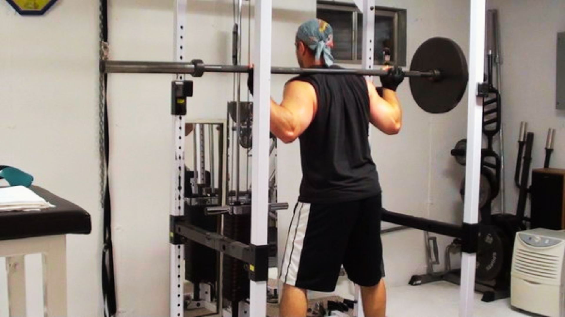 Man in a gym, wearing a bandana and gloves, performs a squat with a barbell on his shoulders, showing concentration and strength.