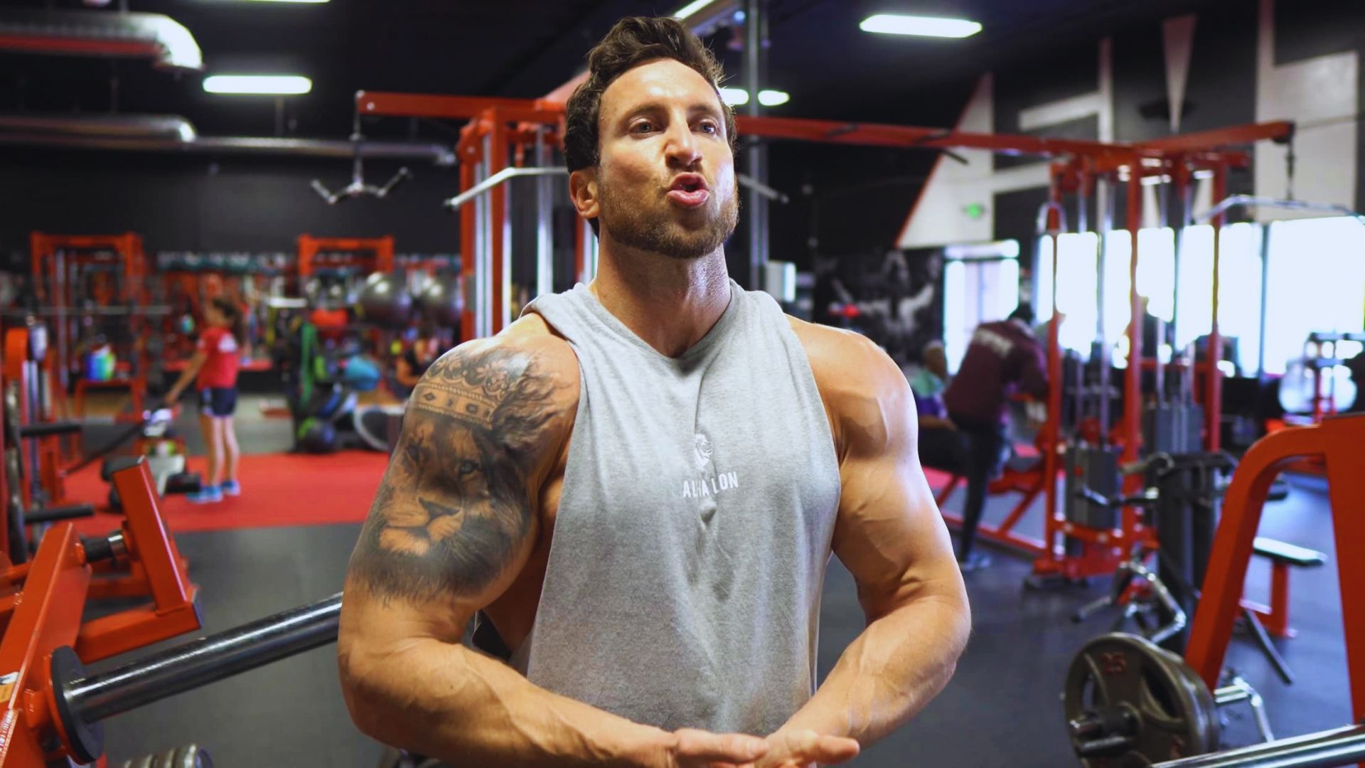 A tattooed man stands confidently in a gym, surrounded by fitness equipment and weights.