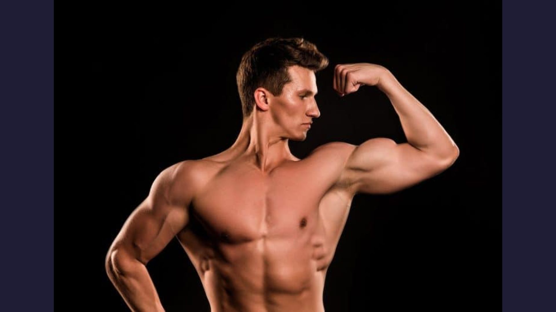 A man flexes his muscles against a solid black background, showcasing strength and fitness.
