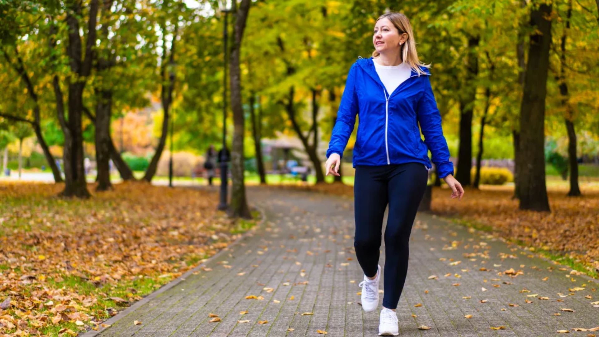 A woman in a blue jacket and black pants walks through a park, surrounded by greenery and trees.