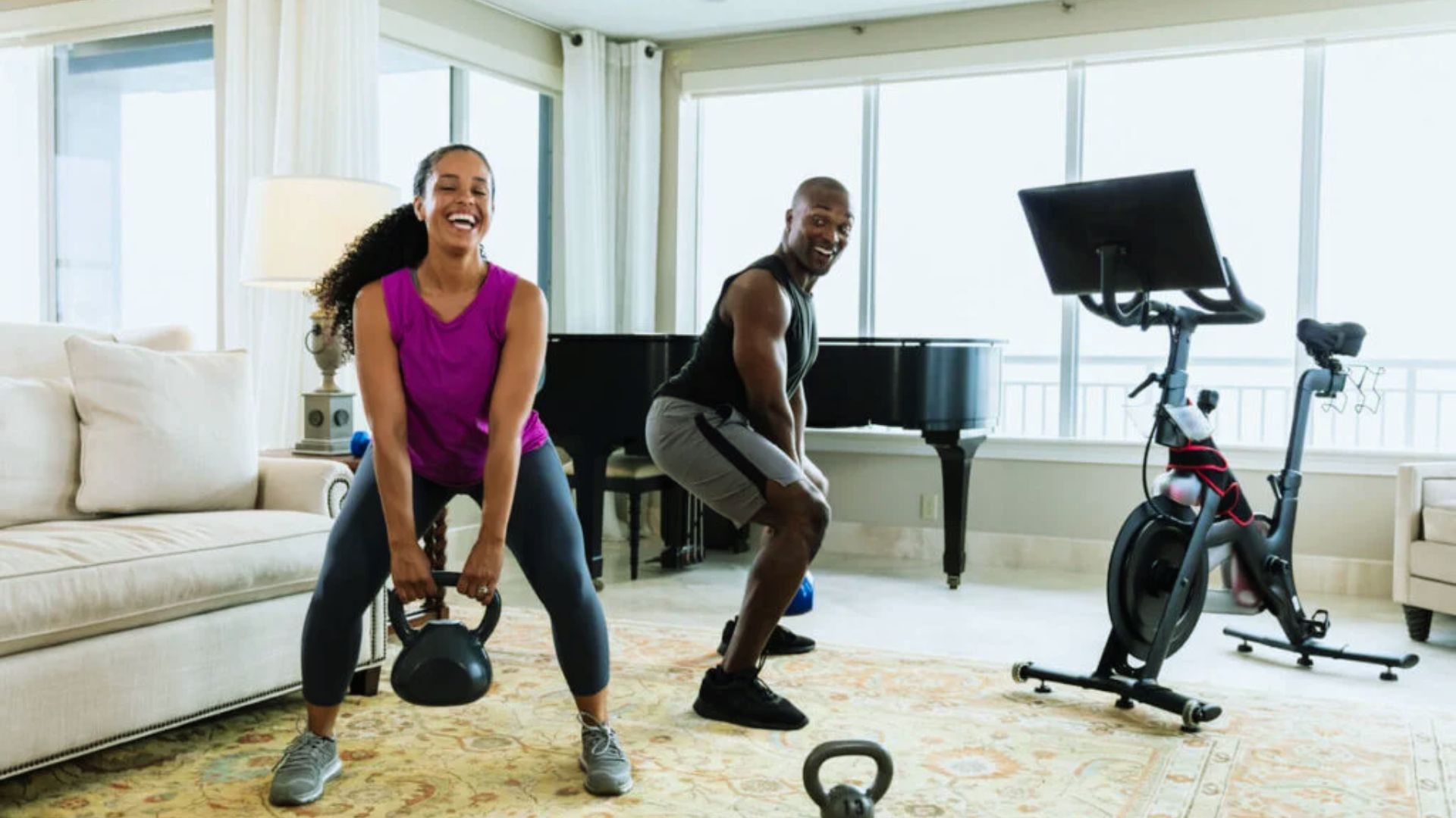 A joyful couple exercises in a bright living room. The woman lifts a kettlebell, while the man squats beside a stationary bike. A grand piano is in the background.