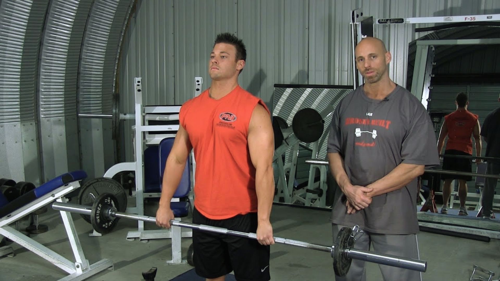 A man in a red tank top holds a barbell, focused and determined, with a coach in a gray shirt standing beside him in a well-equipped gym.