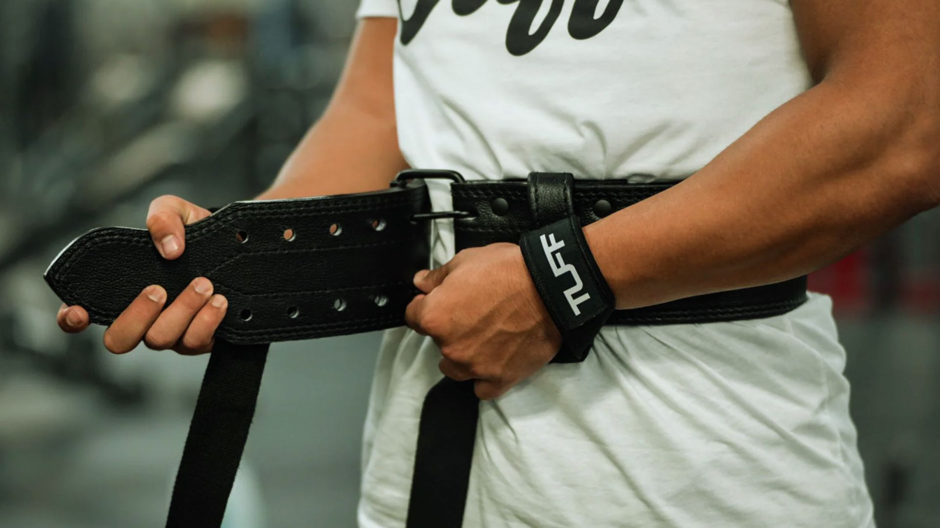 A person adjusts a black leather lifting belt, with "TUFF" on a wrist strap, over a white shirt. The gym background is blurred, conveying focus.