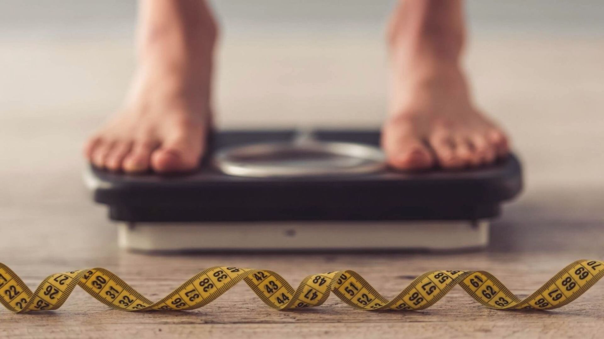 Feet on a bathroom scale with a yellow measuring tape in the foreground. The image conveys themes of weight measurement and health awareness.